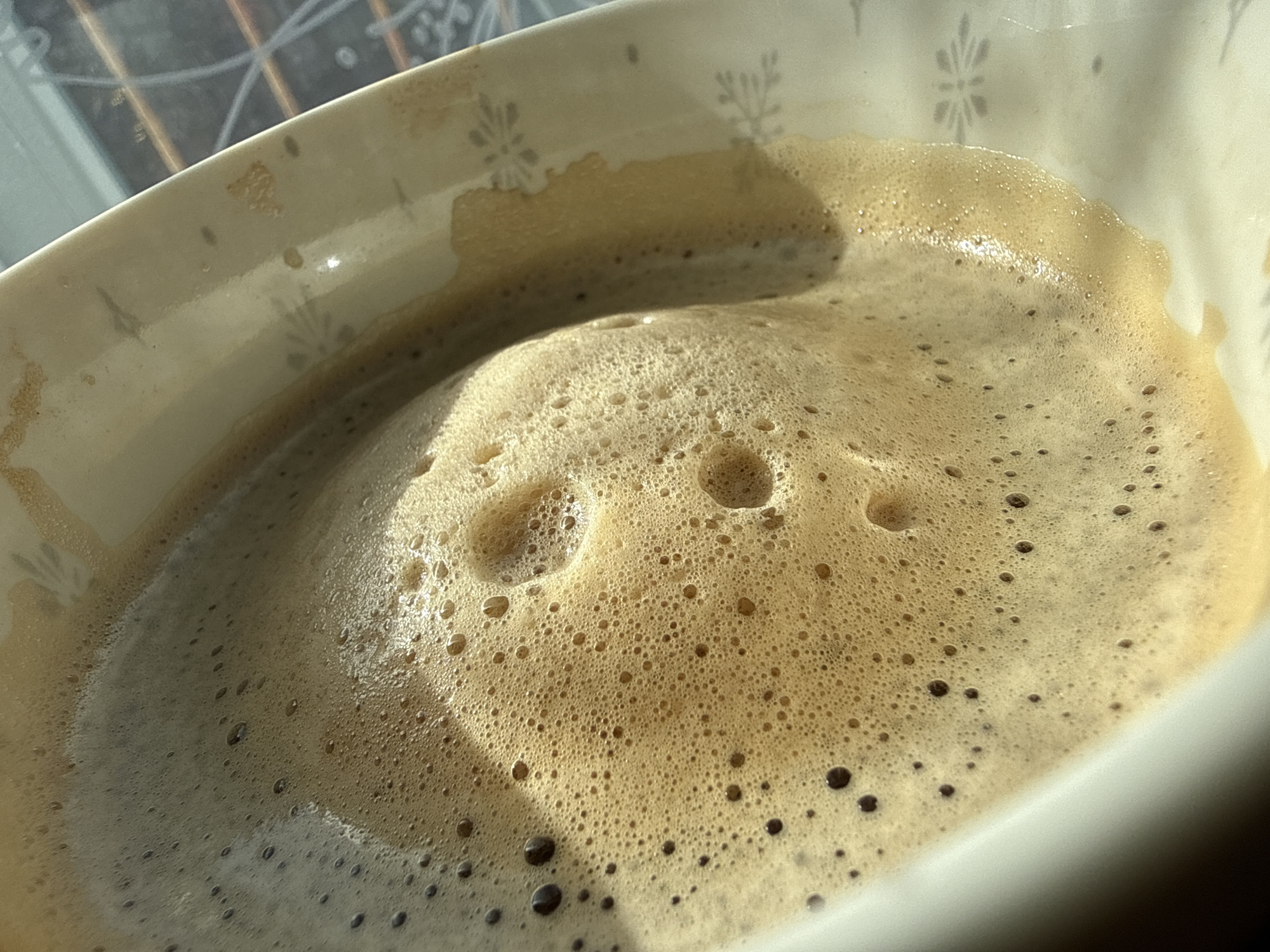 The image shows a close-up of a coffee drink with a frothy surface in a white bowl. The bowl has a pattern of small, grey floral designs. The coffee is light brown with a foam layer of varying bubble sizes. Some bubbles are dark brown. Light from a window illuminates the scene from the upper left, casting a shadow on the lower right of the bowl. A window or window frame is visible in the upper left background. There is no indication of the location of Varna, Bulgaria in the image. There are no people present in the image.