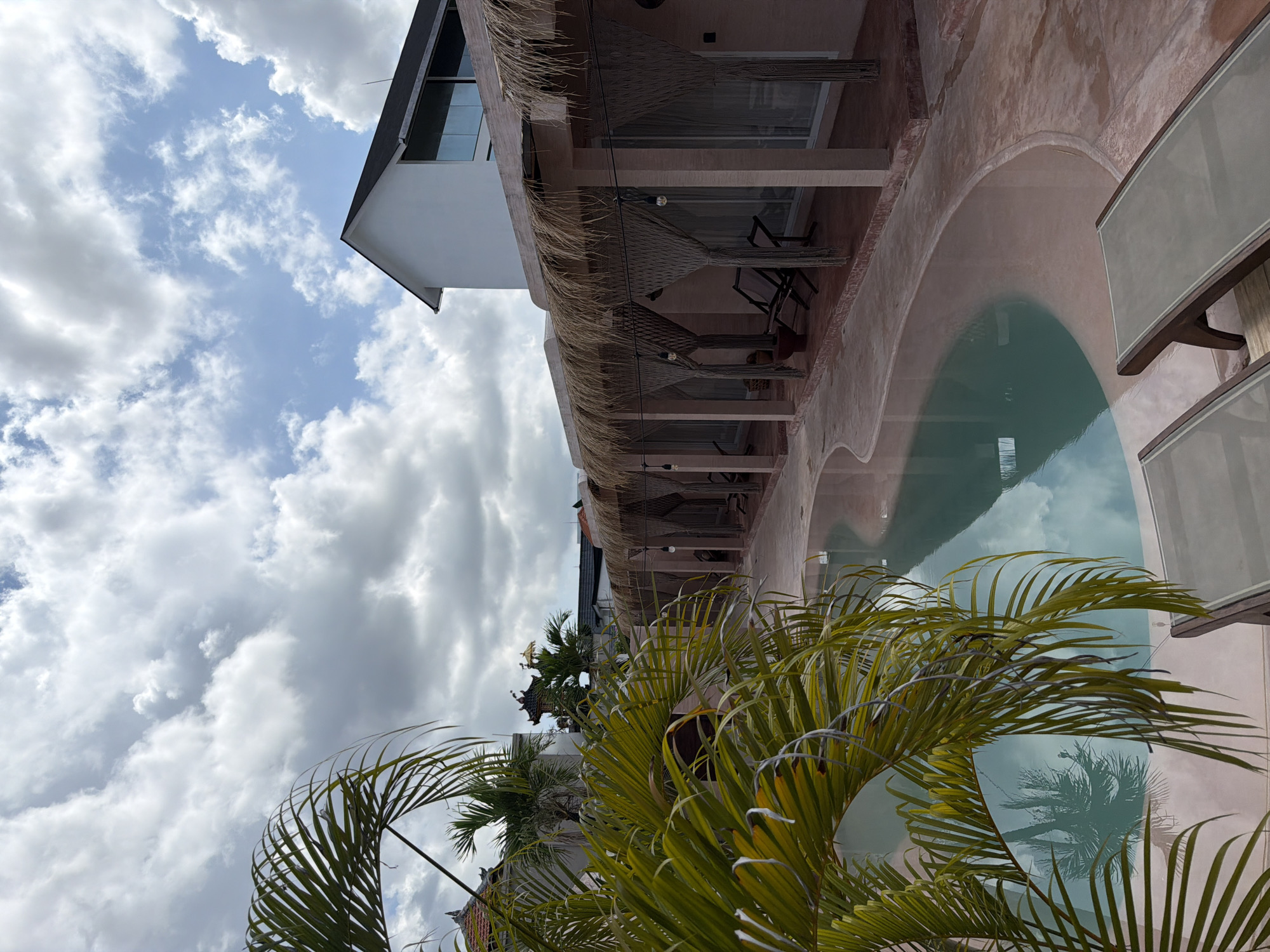 The image captures a view in North Kuta, Indonesia, showcasing a multi-level building with thatched roofs, balconies equipped with hammocks, and an adjacent pool reflecting the sky. In the foreground, lush palm foliage partially obscures the view, adding to the tropical ambiance. The building appears to be a resort or hotel, situated in a location that emphasizes the natural landscape and relaxing atmosphere. The scene is set under a partly cloudy sky, suggesting a clear or recently clear weather.