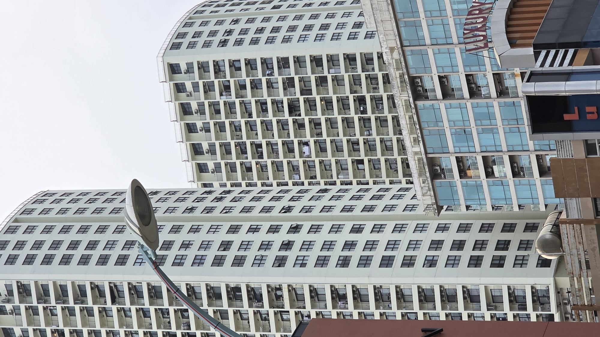 A vertical photograph captures a dense urban landscape featuring multiple high-rise buildings and prominent streetlights against an overcast sky.
In the left and foreground of the image, a large, light-colored building with a gentle curve in its facade dominates. This building is characterized by numerous repetitive balconies, each typically equipped with an air conditioning unit. On several balconies, clothes are seen hanging out to dry, indicating residential use. A single person in a white shirt is visible on one of the balconies near the building's center. The upper section of this building displays many small, square windows.
To the right, a more modern, taller building rises, featuring a facade primarily composed of blue-tinted glass curtain walls. A large red sign is affixed to its exterior, clearly displaying the word "LUXURY" in capital letters. Below this, another sign with dark, reflective surfaces shows partial text that appears to be "LB".
Two streetlights with long, curved poles and sleek, silver-grey fixtures are noticeable. One extends from the lower left, curving towards the center, while the top of another is partially visible on the right side of the image. The sky overhead is uniformly bright and overcast, contributing to diffused lighting across the scene, suggesting daytime.
The image portrays a busy city environment, a common sight in urban areas like Nha Trang Ward, Vietnam, where modern architecture blends residential and commercial functions.