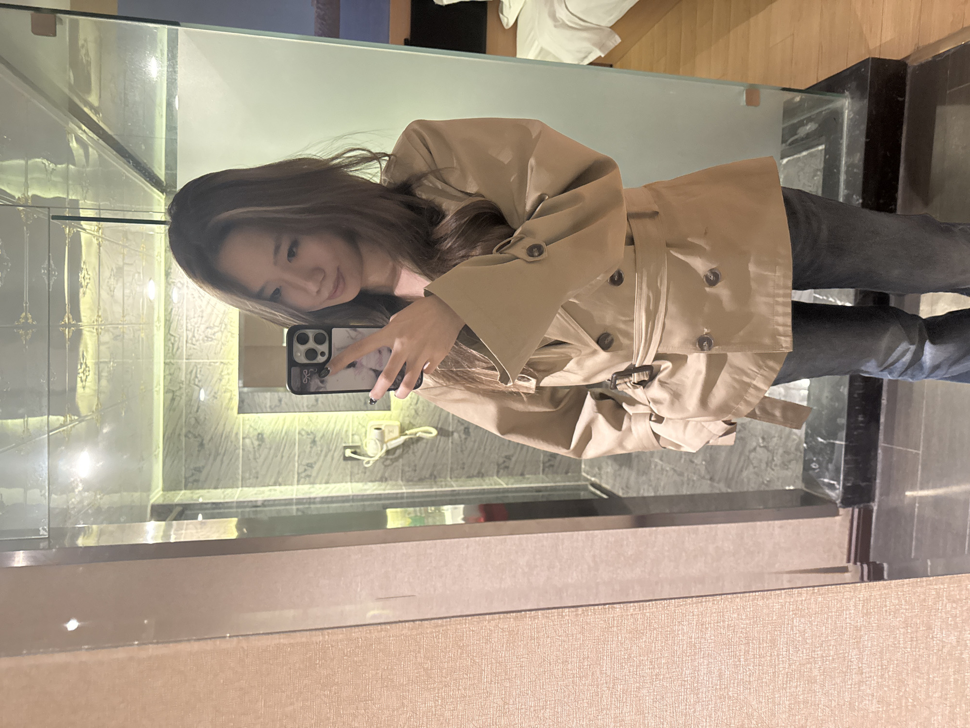 A young East Asian woman is captured in a mirror selfie, holding a dark-colored smartphone with a light-patterned case featuring a cat design. She is wearing a stylish light brown or beige trench coat, cinched at the waist with a belt, and dark jeans. Her long, light brown hair with highlights frames her face as she looks towards the phone with a subtle, pleasant expression. Her left hand, adorned with dark nail polish, holds the phone firmly.
The setting appears to be a modern hotel room, likely in Ganzhou, China. The mirror reflects a well-lit interior, showcasing contemporary design elements. Behind the woman, a tiled wall with a grey marble pattern and subtle decorative accents is visible, suggesting a bathroom area. A dark-colored counter or vanity top runs beneath the mirror, and a clear glass partition, possibly for a shower or to separate the bathroom from the sleeping area, can be seen. On the tiled wall, a white electrical outlet or fixture with a cord is visible.
In the reflected background, elements of the wider hotel room are discernible. A light brown wooden floor is visible, along with white bed linen or towels, confirming the hotel room environment. The foreground shows a textured, light brown paneling or wall surface, likely part of the mirror frame or an adjacent wall. The overall lighting is artificial and bright, typical of an indoor hotel setting. The activity depicted is a casual moment of self-photography. There is no visible text.