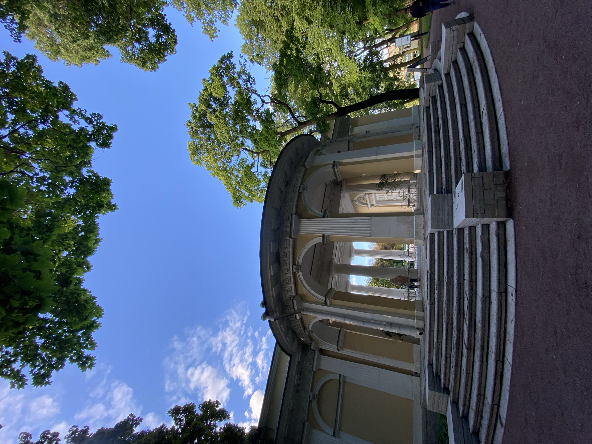 This is a low-angle, full shot of a neoclassical rotunda building in St. Petersburg, Russia. The building has a pale yellow facade, with arched openings supported by white columns. A wide, multi-tiered stone staircase leads up to the rotunda's entrance. Lush green trees with visible leaves surround the building, with some branches extending into the bright blue sky. White, wispy clouds are scattered across the sky. A single person, wearing a hooded jacket, stands at the top of the stairs, facing away from the camera. To the right of the building, a person in a dark jacket is walking away from the camera on a gravel path. The environment appears to be a park or garden setting, with the ground covered in dark gravel.