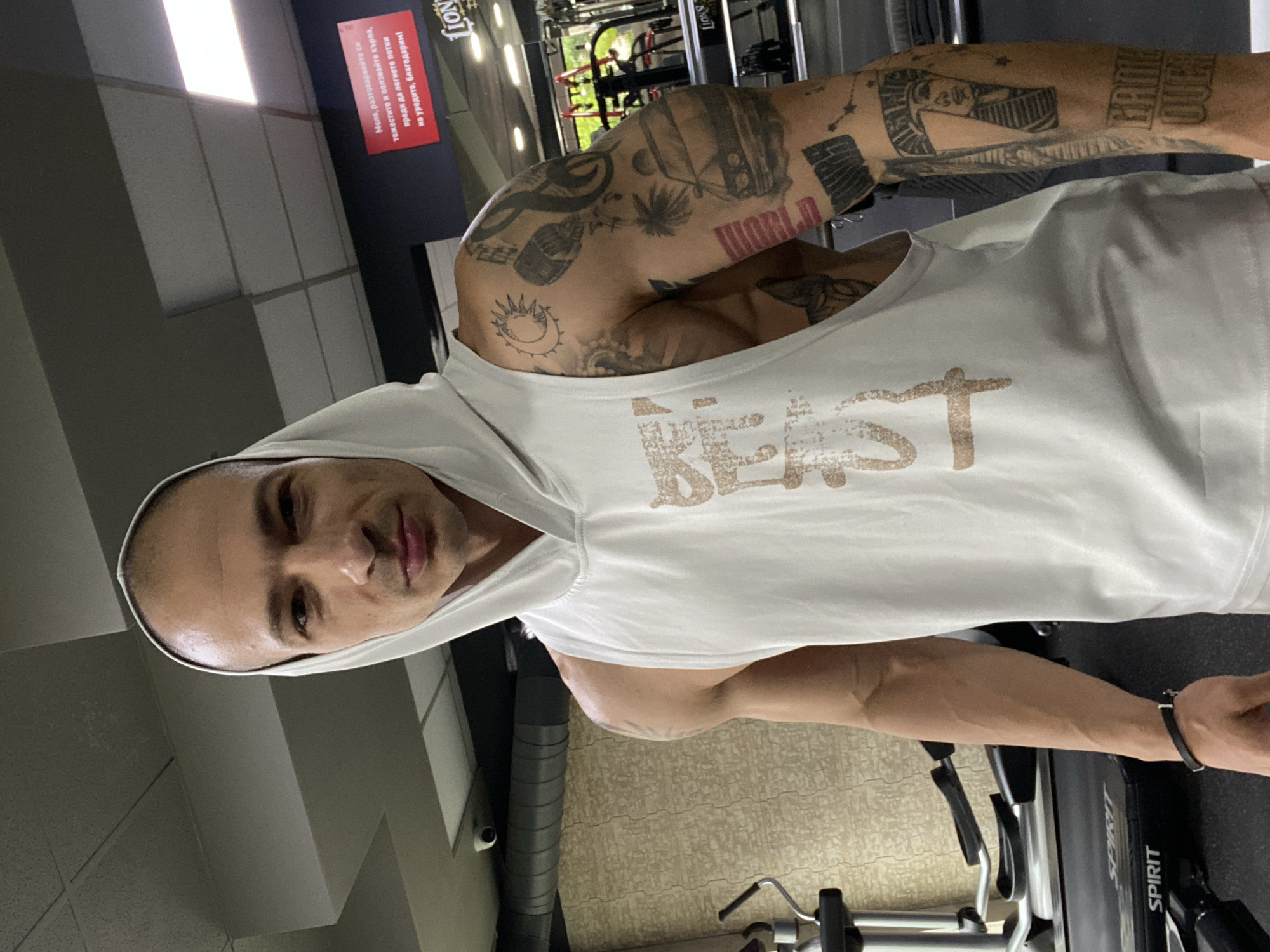 A man stands indoors wearing a white hooded tank top with the word "BEAST" printed on it in gold-brown letters. He is in what appears to be a gym, standing next to a "Spirit" treadmill. He has tattoos covering most of his arms. A red sign hanging on the wall in Bulgarian reads: "Моля, разтоварвайте си тежестите и ползвайте кърпа, преди да легнете потни на уредите, благодарим!" and the words "LION", and possibly "LIONS" below. The location is presumably a gym in Pleven, Bulgaria.