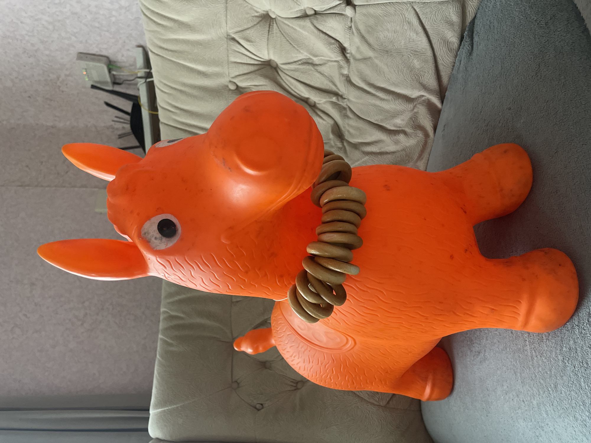 An orange, toy horse with a string of wood bagel pieces around its neck rests on a gray sofa. The sofa is partially situated on a light beige, tufted couch. The location is unspecified and there is no evidence to determine the city or country.