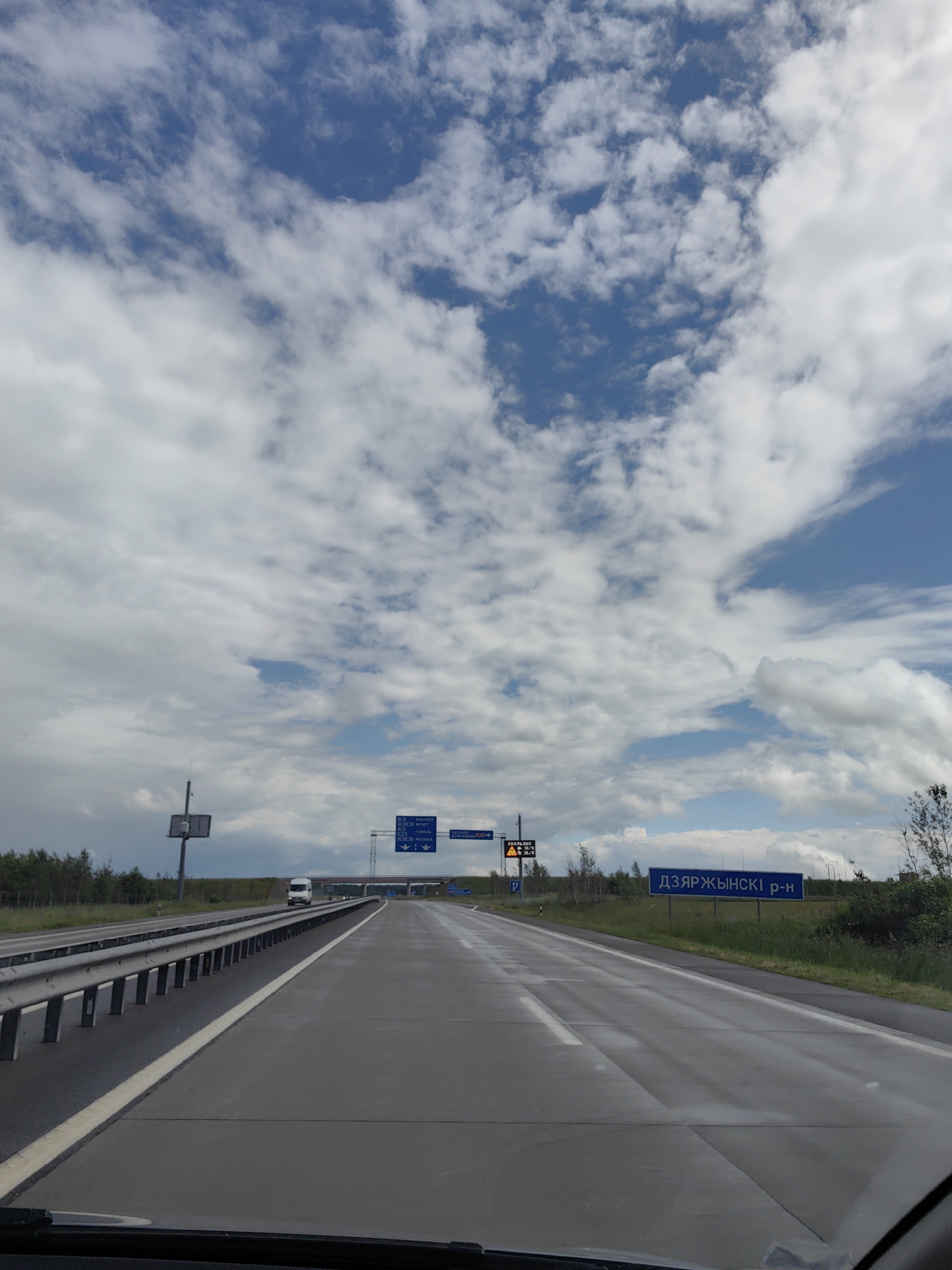 The image shows a highway view in Путчынскі сельскі Савет, Belarus. The sky is filled with white, fluffy clouds against a blue backdrop. The highway is paved and wet, with white lines marking the lanes. A metal guardrail runs along the left side of the road. In the distance, a white van is driving on the highway. There are several road signs visible. One blue sign on the right side of the road reads "ДЗЯРЖЫНСКІ р-н". Another blue sign on the left side shows directions to various cities, including "МИНСК", "ГРОДНО", "ВЯЗЬМА", and "МОСКВА". There are also some smaller electronic signs on the left indicating traffic information. Trees and vegetation line the sides of the highway.