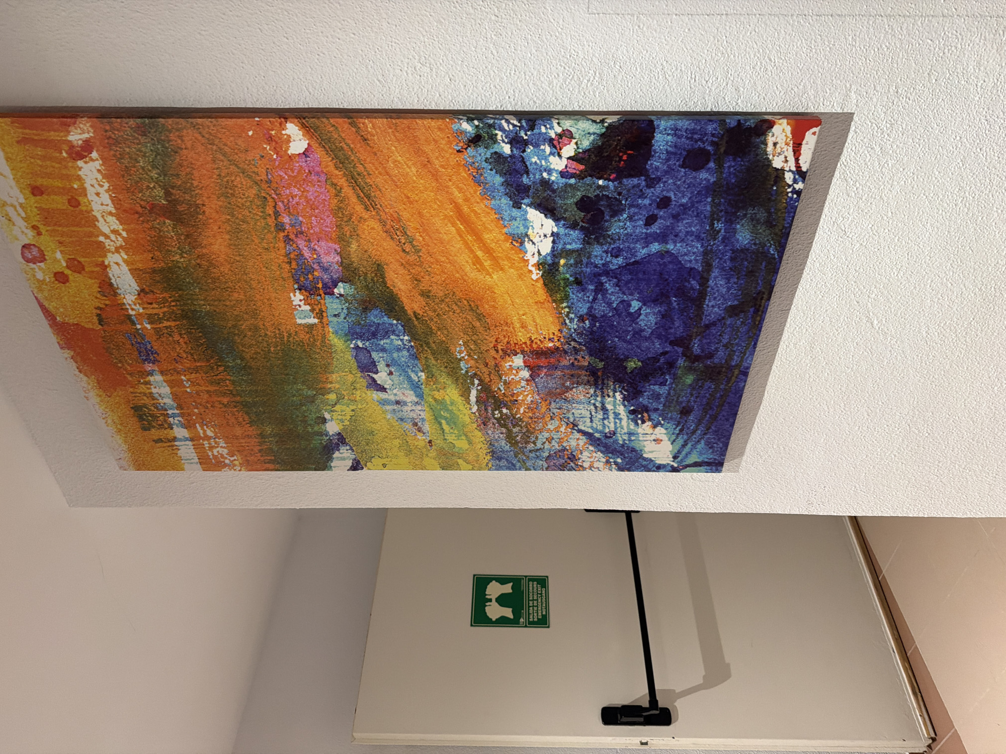 The image shows a painting with abstract brushstrokes in shades of orange, blue, and yellow, mounted on a white textured ceiling. Below the painting, a closed white door is visible. On the door, there is a green and white emergency exit sign with text in Spanish and English that reads "SALIDA DE SOCORRO" and "EMERGENCY EXIT". A black pull handle is attached to the door, and a thin black cord extends upwards from it. The lighting suggests an indoor setting. The overall scene appears to be in a public space, possibly a hotel or a commercial establishment in Sant Antoni De Portmany, Spain.