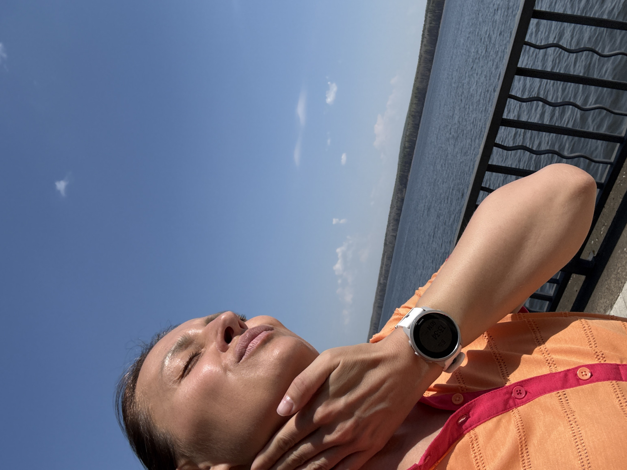 A woman is photographed in an outdoor setting, likely in Chaikovsky, Russia, judging by the water body and vegetation. The view is angled upwards towards her face. Her eyes are closed, and she is holding her own neck/throat with her hand. She is wearing an orange shirt with pink trim and buttons. A white smartwatch is visible on her left wrist, displaying the time (13:54). The background includes a metal railing and a body of water, possibly a river or lake, with a treeline visible on the opposite shore. The sky above is blue with sparse clouds. The overall lighting indicates it is a sunny day.
