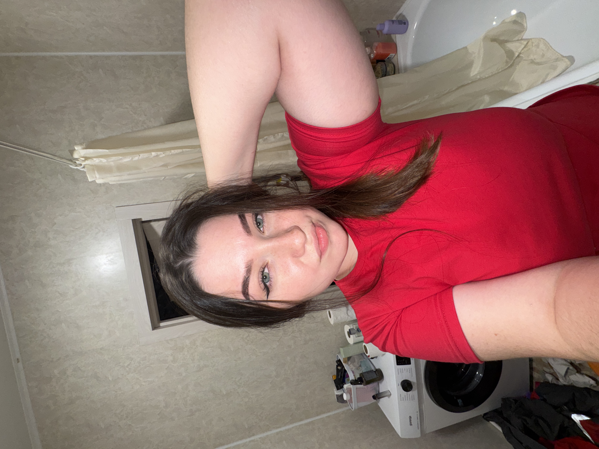 This image, taken in Kitsjanovo, Russia, captures a young woman posing for a selfie in a domestic bathroom. The photo is oriented 90 degrees clockwise from a standard portrait view.
The main subject is a woman with long, dark brown hair, striking blue-green eyes, and full lips, who is looking directly at the camera with a calm and slightly smiling expression. She is wearing a bright red short-sleeved top. Her bent legs are visible in the upper right quadrant of the image, suggesting she might be lying in a bathtub or on the bathroom floor.
The bathroom features light-colored, subtly patterned tiled walls. To the right, a white bathtub contains various plastic bottles of toiletries on its edge, next to a beige shower curtain that is partially drawn. Below the bathtub, a white front-loading washing machine, branded "Gorenje", is visible. On top of the washing machine, several rolls of toilet paper are stacked, alongside a small container holding miscellaneous items. A pile of dark clothes is situated next to the appliance. A rectangular vent or window frame is visible on the wall to the left of the woman's head. The lighting appears to be artificial, typical of an indoor bathroom setting.