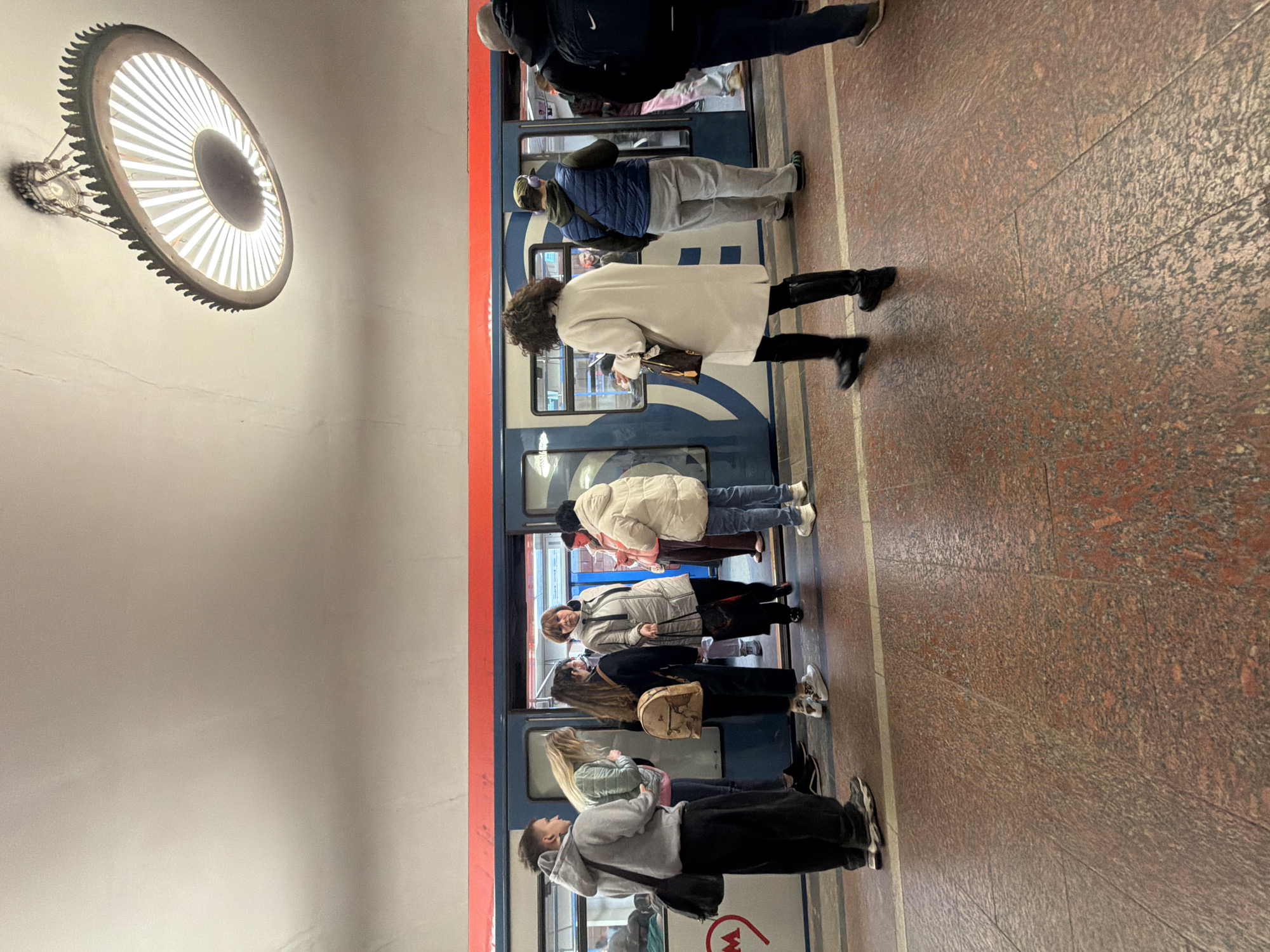 The image shows a subway platform in Moscow, Russia. People are waiting to board a train. The train doors are open, and some passengers are visible inside. The platform has a red granite floor and white walls. A large, circular light fixture hangs from the ceiling. The train is painted in blue and white stripes. Most people on the platform are wearing winter clothing and face masks.
