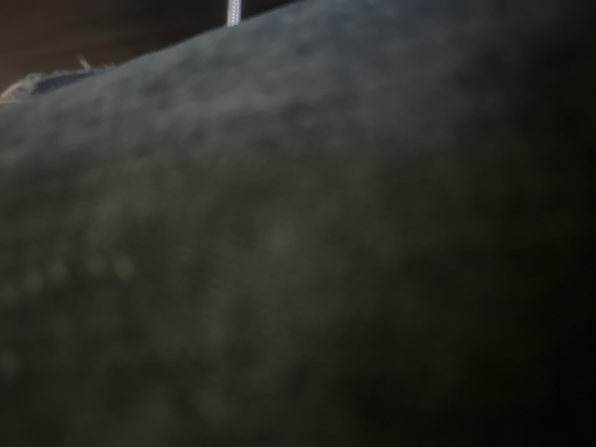 An abstract, low-light photograph taken in Casablanca, Morocco, depicts a dark, textured surface occupying most of the frame. The surface, likely a fabric such as denim or suede, appears charcoal grey with subtle hints of dark olive green, exhibiting a soft, mottled texture due to extreme blur and dim illumination. Faint, lighter, circular or irregular blurs are dispersed across its surface, possibly indicating wear, minor reflections, or smudges. On the right side, a distinct, slightly lighter vertical edge of the fabric is visible, showing frayed fibers, consistent with a seam or a worn garment hem. Behind this edge, an indistinct, dark brownish surface, possibly wood, recedes into shadow. In the upper right corner, a thin, light-colored, possibly braided cord or cable is faintly discernible against the dark background. No individuals, specific actions, or other identifiable objects are present. The overall environment is characterized by extreme dimness and a complete lack of sharp focus.
