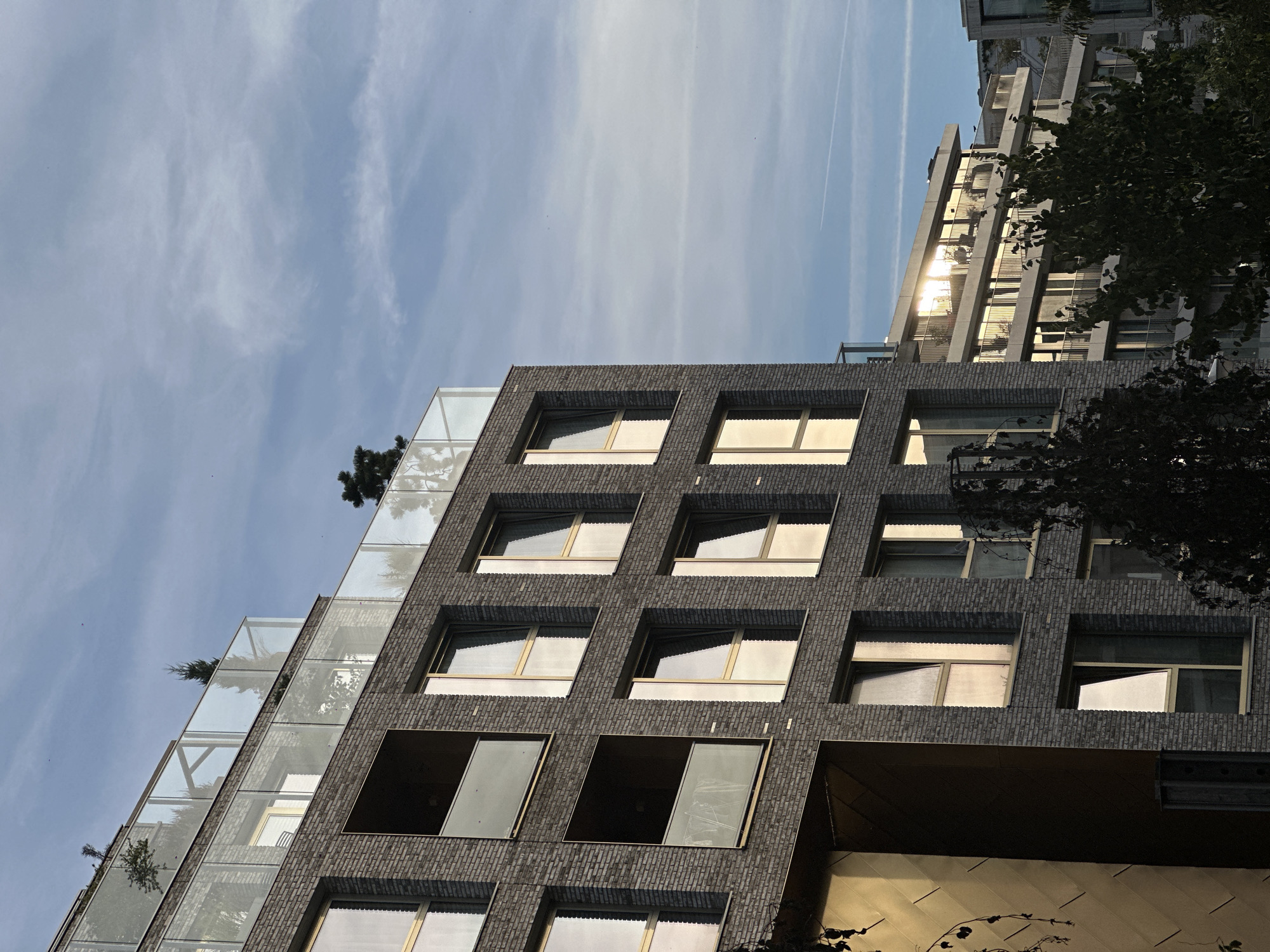 This image depicts the exterior of a modern apartment building in Amsterdam, Netherlands, viewed from a low angle against a clear, pale blue sky with wispy clouds and contrails. The building is constructed with dark grey brick and features multiple floors with large windows and balconies. The windows are framed in gold and reflect the warm glow of the setting sun. Some windows are open, and balcony railings are made of glass. Green trees are visible in the foreground and around the building, and a rooftop garden with small trees and glass enclosures is present on the top floor. Another building with similar modern architecture, featuring balconies and reflective windows, is visible in the background to the right. The overall scene is tranquil and captures the architectural style of contemporary urban development in Amsterdam.