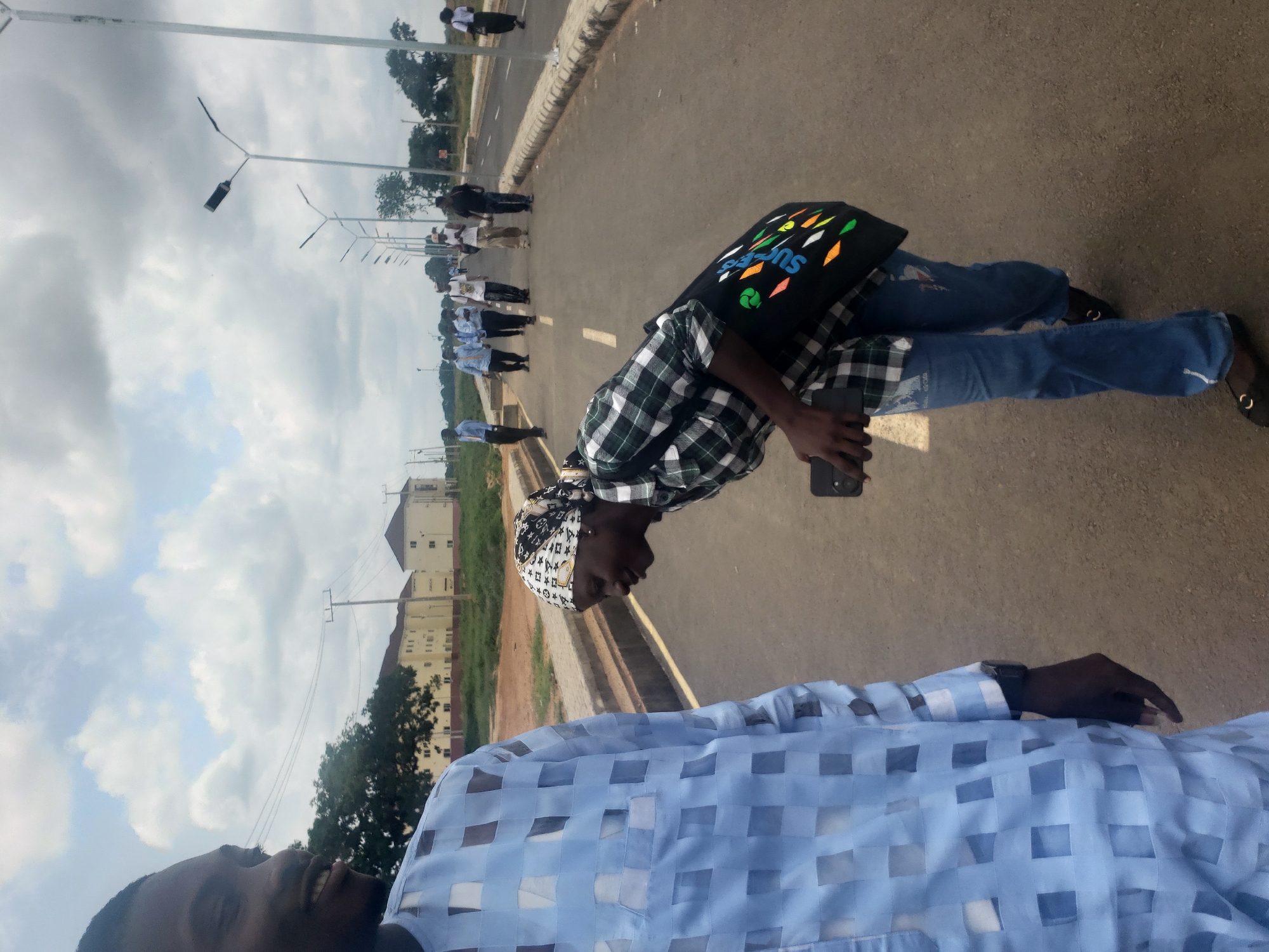 A medium shot captures a group of people on a paved road with streetlights lining the right side. In the foreground, to the left, a person wearing a light blue, patterned garment is partially visible, looking towards the left. To the right of this individual, a woman wearing a green and white checkered shirt and a headscarf is bent over, holding a dark-colored smartphone. She is also carrying a black tote bag adorned with colorful geometric shapes and text. The road continues into the background, where several other individuals are walking away from the camera, some in formal attire. Buildings with light-colored facades and multiple stories are visible in the distance on the left side of the road, set against a backdrop of trees and a cloudy sky. The overall setting appears to be an urban or semi-urban environment in Gumel, Nigeria.