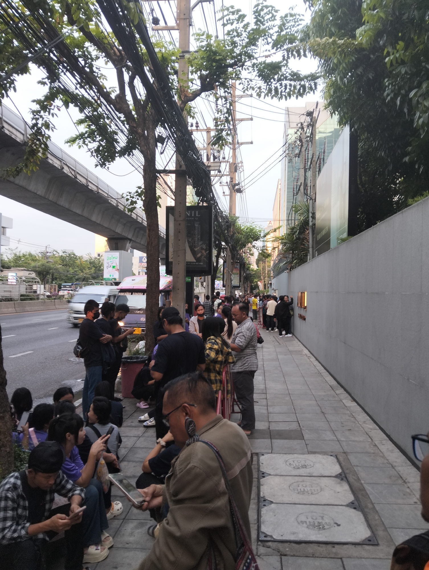 The image shows a queue of people on a sidewalk in Bangkok, Thailand. The people are waiting in front of a building with a long, gray wall. Some are sitting on the ground while others stand. Many are looking at or using their mobile phones. The queue stretches along the sidewalk, next to a road with vehicle traffic. Above the road is an elevated train track. Trees and utility poles with dense networks of wires line the street. Signs are visible in Thai script, indicating the location is Bangkok. The scene depicts a casual, everyday activity within an urban environment.