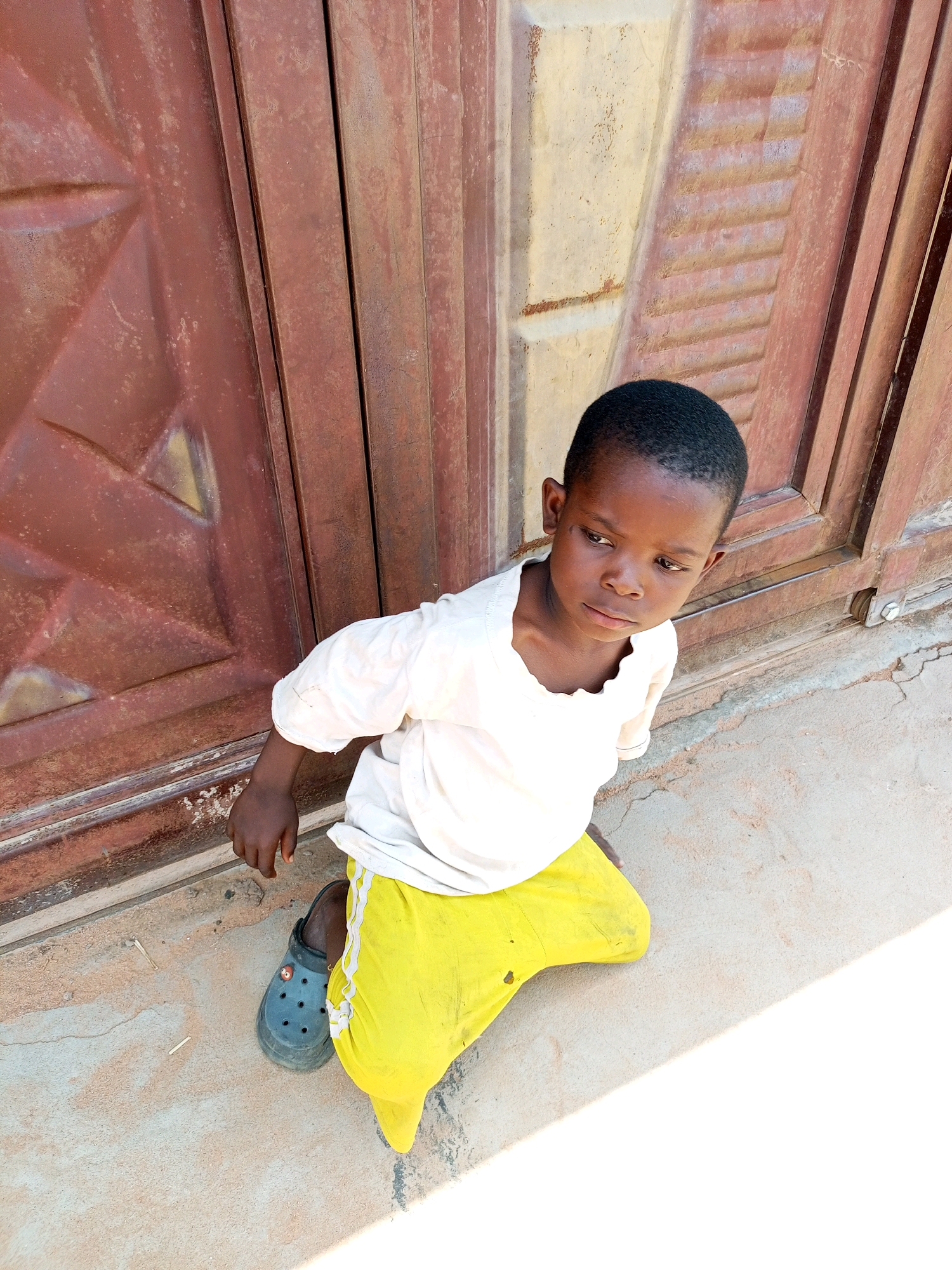 A young Black child kneels on a concrete surface in front of a large, weathered, reddish-brown metal door. The child is wearing a short-sleeved white shirt and bright yellow pants. A black Croc-style shoe is visible on the child's left foot. The door appears to be a roll-up or security door, with horizontal corrugated panels on the right side and a diamond pattern on the left. The concrete ground is dusty and cracked, with a few small debris items scattered on it. Sunlight casts a sharp shadow across the lower right portion of the image. The child's gaze is directed downwards and to the right. The scene is likely outdoors, possibly in a commercial or residential area in Tempure, Nigeria.