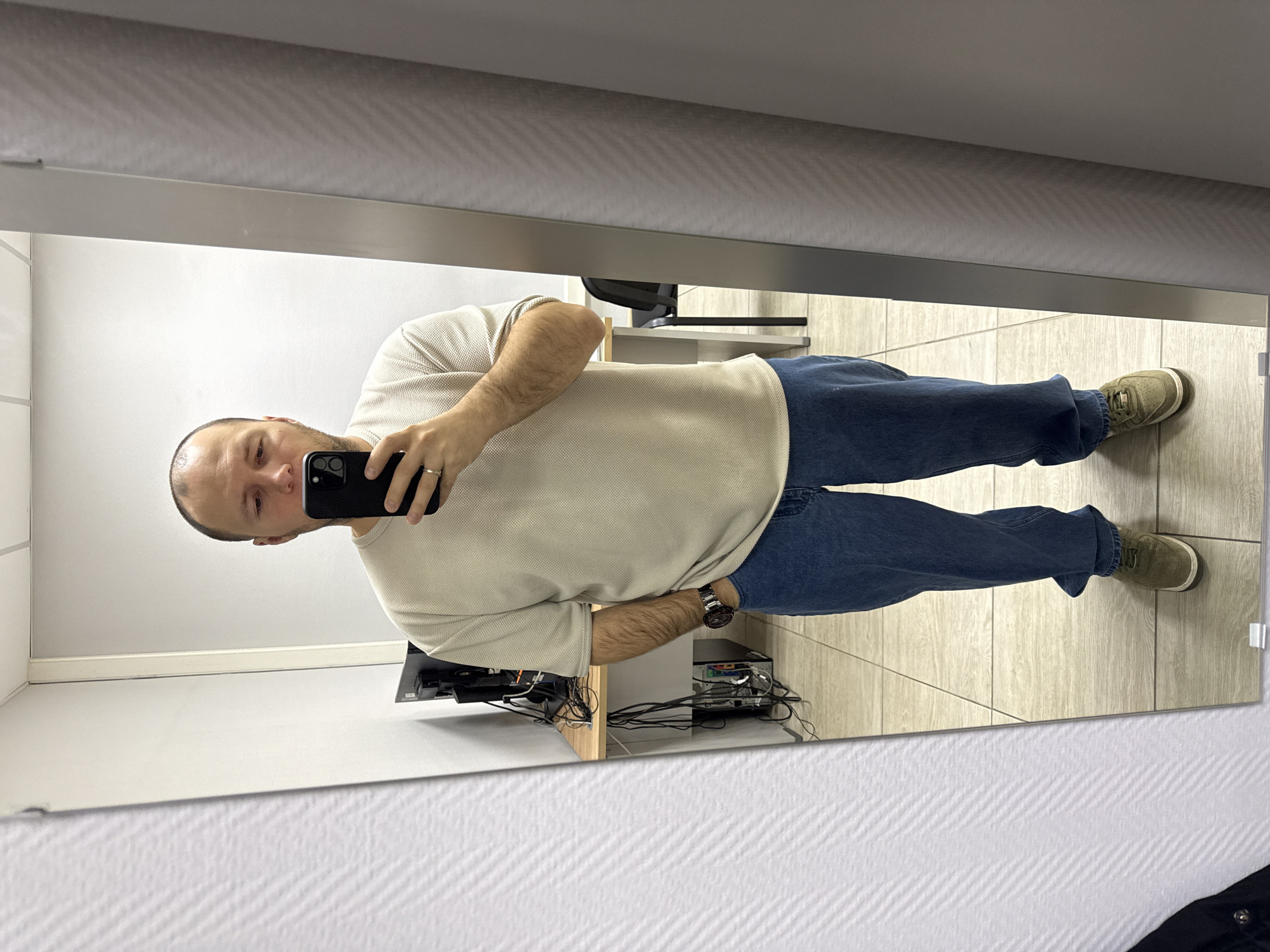 A reflection in a mirror shows a man taking a selfie. He is wearing a light-colored, short-sleeved shirt, blue jeans, and green shoes. He is holding a black smartphone. A wristwatch is visible on his left wrist. The reflection shows a room with light-colored walls and tiled floors. In the background, there's a desk with computer equipment and a chair.
The image does not provide information confirming the location is Krasnoyarsk, Russia.