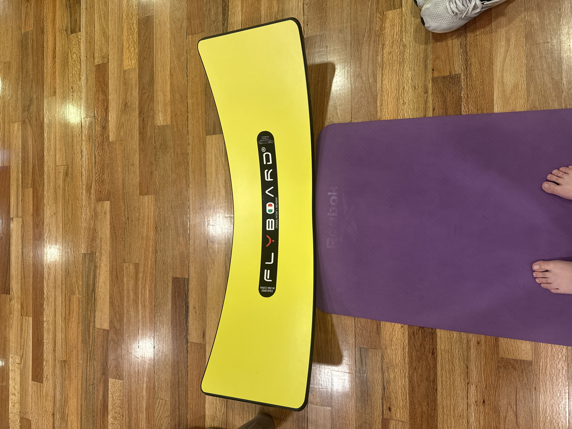 The image shows a yellow Flyboard on a wood floor, with a purple Reebok yoga mat nearby. A pair of white sneakers and a person's bare feet are also visible on the yoga mat. The location appears to be indoors, possibly a home gym or studio. There's no indication of the location being in the city of Puebla, Mexico.