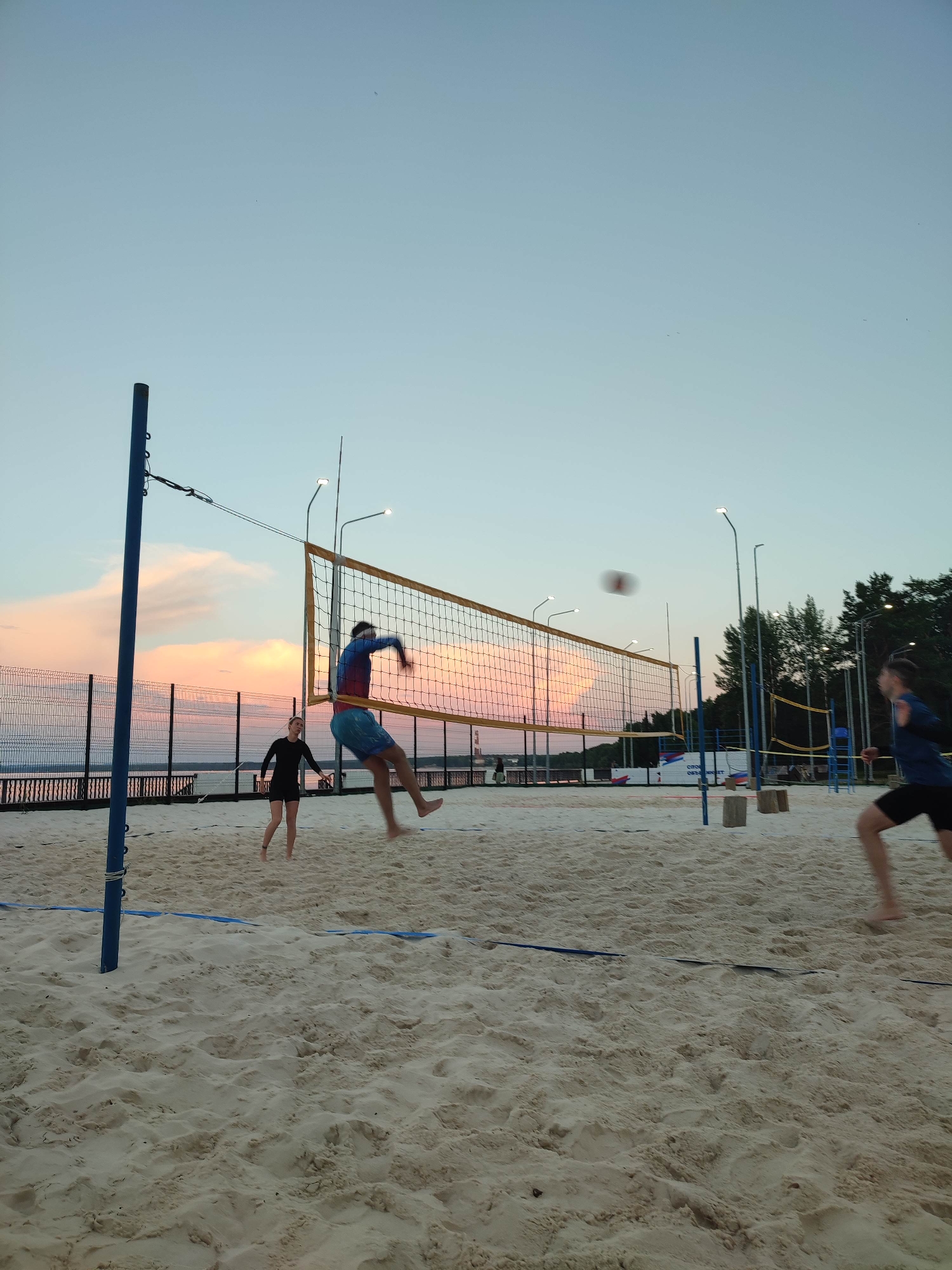 A group of people are playing beach volleyball on a sandy court at dusk. The sky is a soft gradient of blue and pink, with wispy clouds. A volleyball net is stretched across the court, and several players are in motion. One person is jumping high to hit the ball, while another is positioned to receive it. The court is marked with blue lines, and lights are mounted on poles to illuminate the area. In the background, there is a fence, trees, and a body of water. The sand is light in color and shows footprints from previous games.