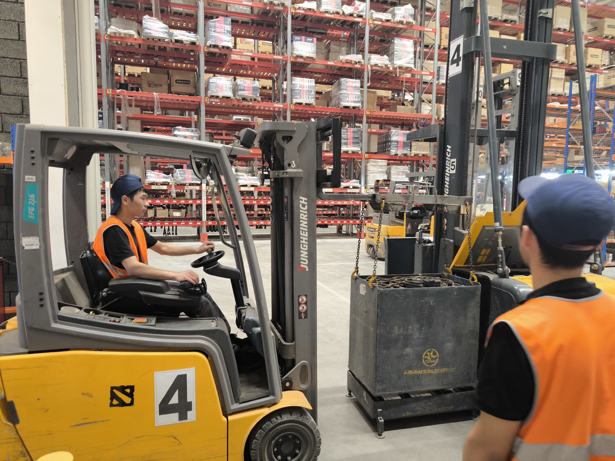 This image shows a warehouse interior, possibly in Domodedovo, Russia, with two forklifts and several rows of high shelving units packed with goods. In the foreground, a yellow forklift is driven by a person wearing an orange safety vest and a blue cap. The forklift has a "4" marked on its side. To the right, another forklift, black and yellow, is partially visible, and in front of it, a large metal bin is being lifted by chains. A second person, also wearing an orange safety vest and a blue cap, stands facing away from the camera, observing the operation. The shelving units are constructed of red metal frames and contain numerous boxes and packaged items, some of which are wrapped in plastic. The overall environment appears to be a commercial logistics or storage facility.
