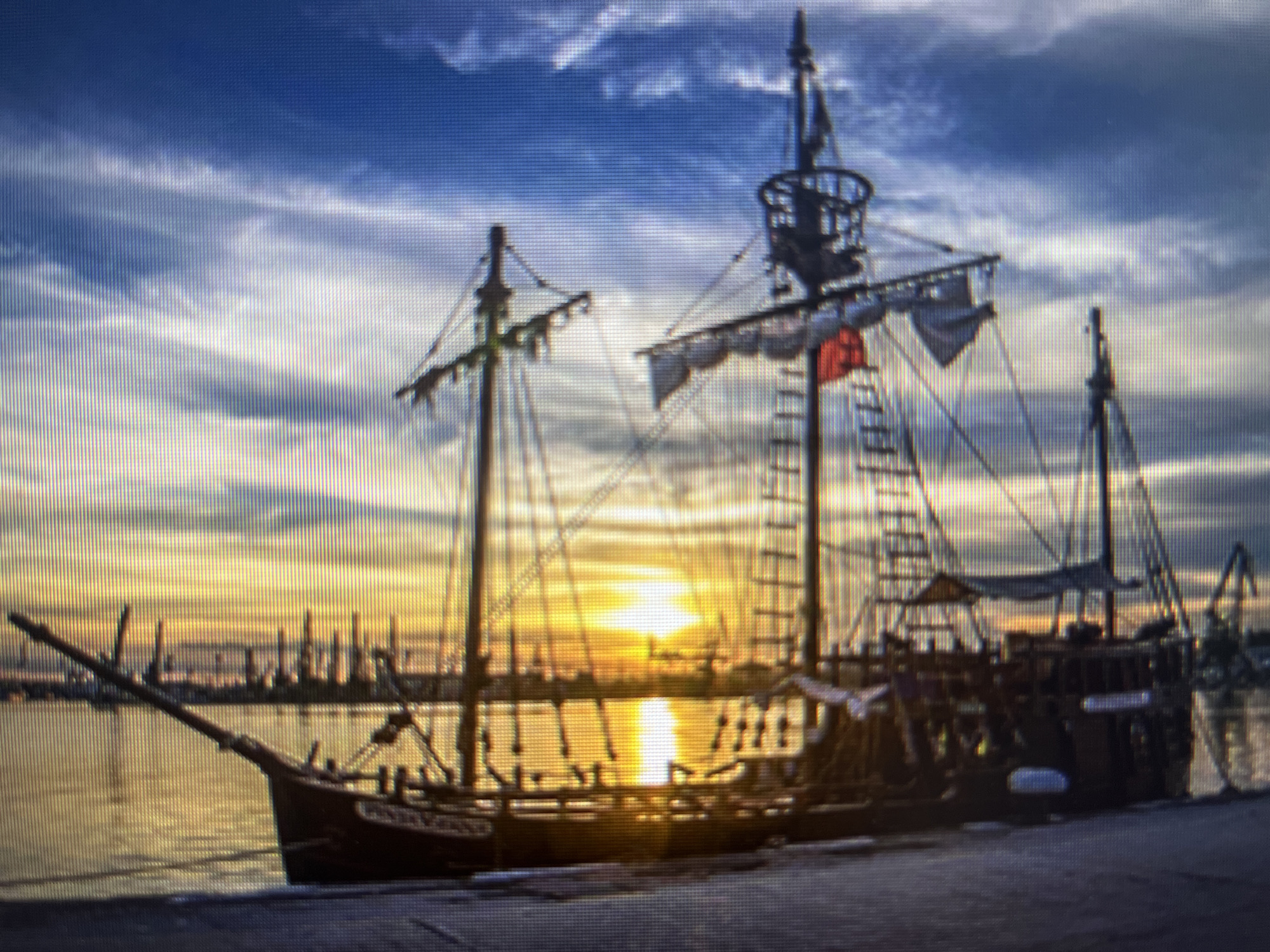 An evocative image depicts a large, multi-masted sailing ship, likely a historical replica, silhouetted against a breathtaking sunset in the harbor of Varna, Bulgaria. The sun, a brilliant golden sphere, is positioned directly behind the ship, casting an intense glow across the water and sky. The sky transitions from a deep blue at the top to vibrant oranges and yellows near the horizon, accented by scattered clouds.
The sailing vessel features prominent masts, intricate rigging, and partially furled sails. A distinct crow's nest can be seen on the tallest mast, and a small red flag is visible. The calm harbor waters beautifully reflect the golden light of the setting sun. In the background, an industrial or city skyline with various structures, including what appear to be cranes, is silhouetted, confirming the port setting. The ship is docked at a dark pier or quay in the foreground. The scene is imbued with a sense of majesty and tranquility, captured at dusk. Due to the resolution, any potential text on the ship is indiscernible.