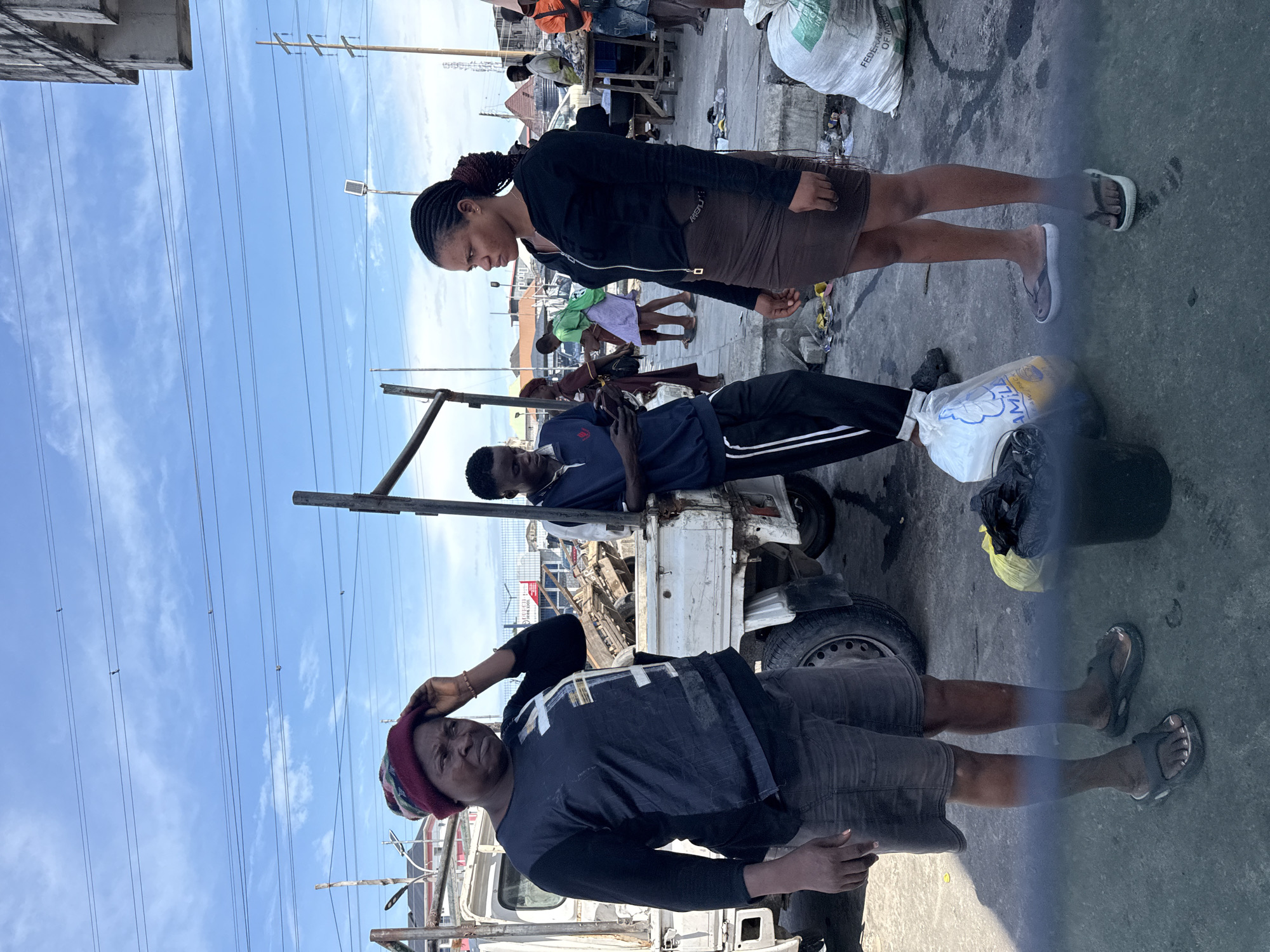 In Igboefon, Nigeria, under a partly cloudy sky, a scene unfolds by a white utility truck. Three individuals are the central focus. A woman on the left, wearing a dark long-sleeved shirt, shorts, and a beanie, stands with her hand on her head, looking to her left. To her right, a young man in a blue polo shirt and black track pants is absorbed in his mobile phone. Further to the right stands a young woman in a black zip-up hoodie and a brown skirt, facing away from the camera and looking to her left.
Behind them, the utility truck is loaded with various items, including what appears to be scrap metal or discarded materials. The ground is paved with concrete and shows signs of wear and debris. In the background, more people are visible, some attending to stalls or engaged in their own activities, suggesting a market or busy roadside area. Power lines crisscross the sky, and a few buildings are visible in the distance. Bags, including a white one labeled "FEDERAL," and a black trash bin are present on the ground near the people. The overall impression is of an outdoor gathering or work area in a Nigerian urban environment.