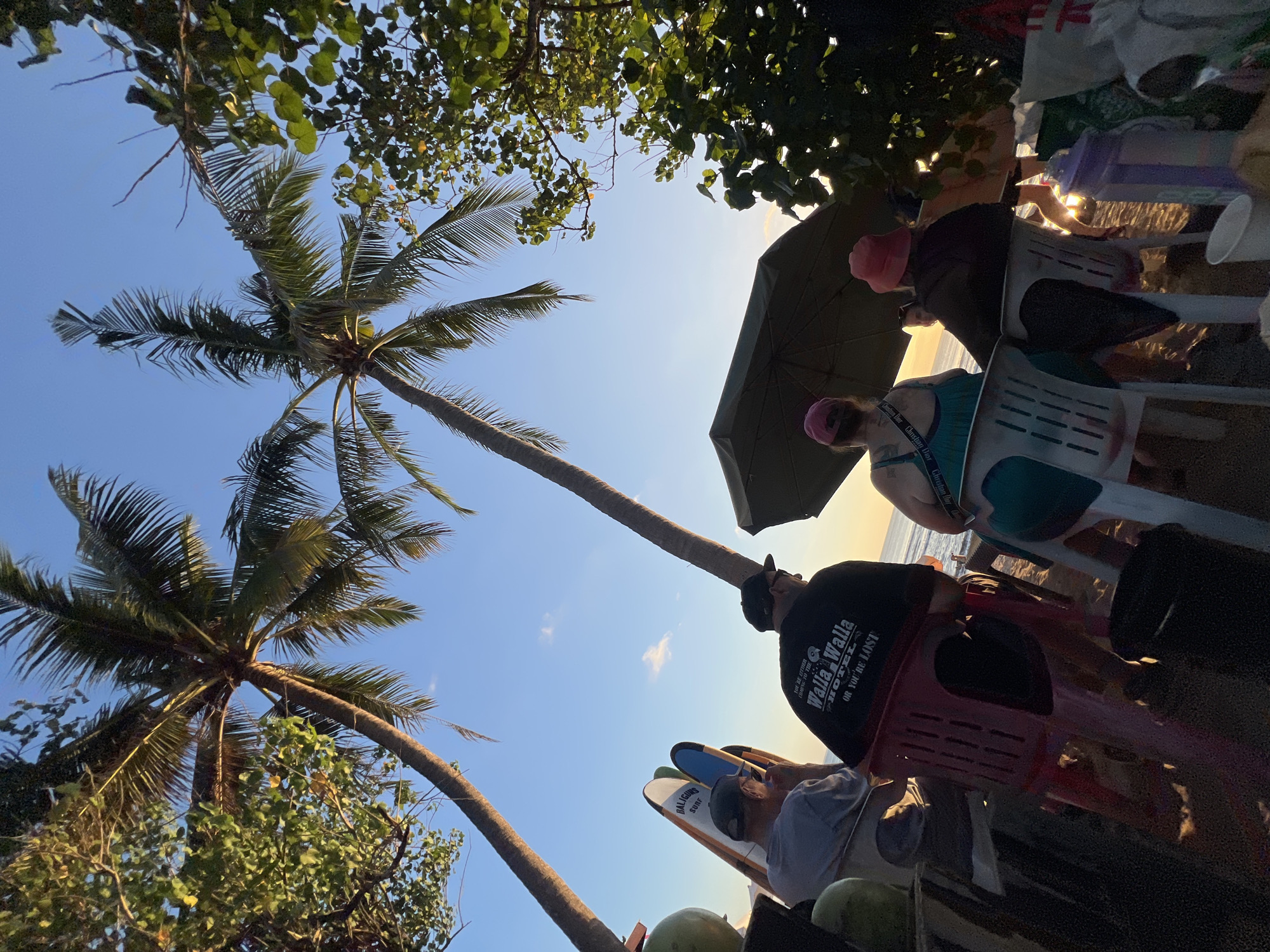 The image depicts a beach scene in Kuta, Indonesia, likely during sunset. Two tall palm trees frame the sky, with their fronds casting shadows on the sand. In the foreground, several people are seated on chairs, facing the ocean. One person in the center wears a black t-shirt with the text "Walla Walla Hotel or You're Lost." Another person to their left is wearing a light gray t-shirt and a baseball cap, and to the right, a person in a teal top is visible. A large umbrella provides shade for some of the individuals. Leaning against a surface to the left are several surfboards, one with the text "BALI GURU SURF." The overall atmosphere suggests a relaxed group enjoying the beach at dusk.