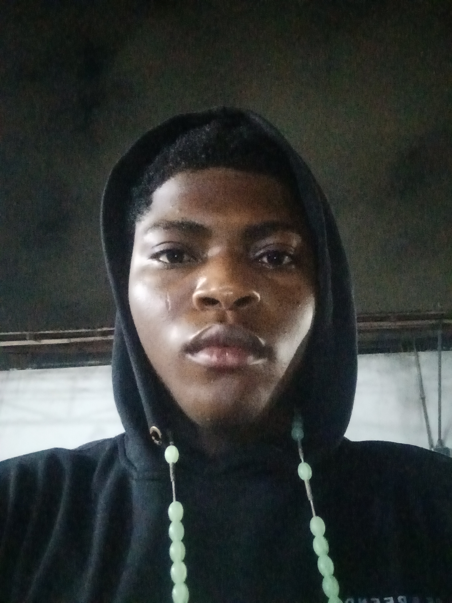 The image shows a close-up, front-facing, self-portrait of a young Black male wearing a black hooded sweatshirt and a string of light green beads. The individual has short, dark hair and appears to be looking directly at the camera. A small, faint scar is visible on the left side of his cheek, near the eye. The background is dark and out of focus, with some lighter, out-of-focus elements suggesting an indoor or covered environment with structures. The lighting appears to be artificial and somewhat dim, casting highlights on the subject's face. There are no other people or significant objects in the frame. The overall setting is nondescript and does not provide specific clues to a location within Ado, Nigeria.