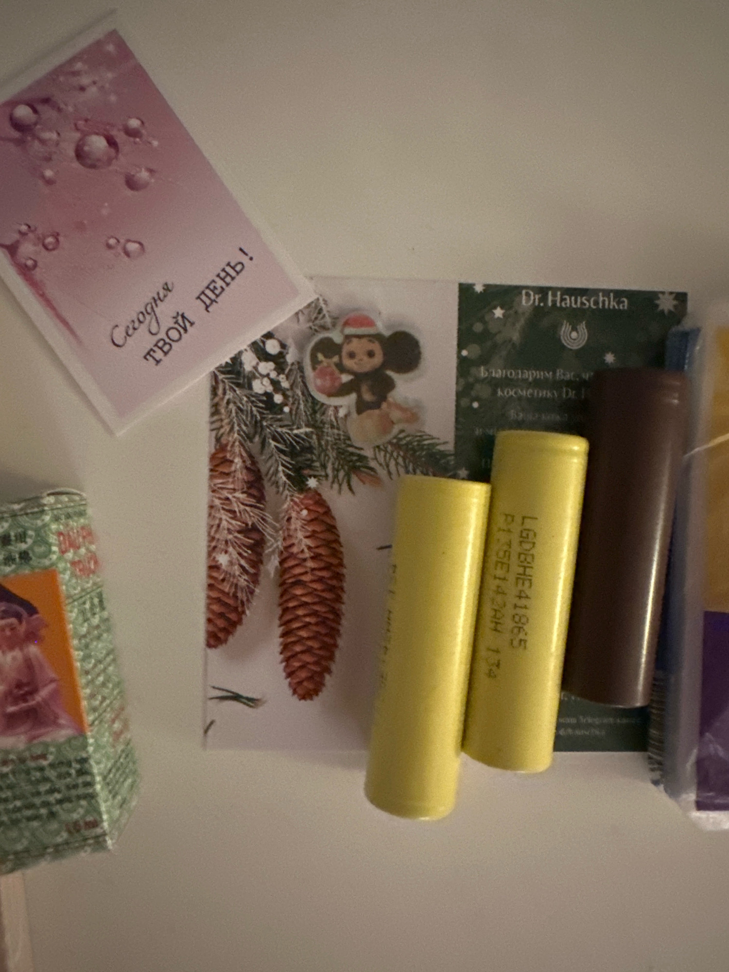 The image depicts a collection of items on a white, textured surface, possibly a wall or a shelf. In the upper left is a pink greeting card with bubbles on it and Russian text that reads "Сегодня твой день!" (Today is your day!). To the right of the card is a small sticker of a cartoon character in a Santa hat. Below the sticker and card, there is a Christmas-themed postcard with pine cones and evergreen branches.
To the right of the Christmas postcard, there is a dark green label with white text that reads "Dr. Hauschka" and some smaller Russian text below it, possibly indicating a cosmetic product. Next to the Dr. Hauschka label, there are three cylindrical objects. The two on the left are yellow, and the one on the right is brown. The yellow cylinders appear to be batteries, with visible text on one of them that reads "LGD BHE 41865 P13SE142AH 174". The brown cylinder is darker and appears to be a lipstick or a similar cosmetic item. Further to the right, a purple and blue object is partially visible.
On the far left of the image, a partially visible green box with intricate patterns and an image of a figure in pink clothing is present. The scene appears to be indoors, with neutral lighting. The image does not provide any specific information about the city of Nevinnomyssk, Russia, or any activities taking place there.