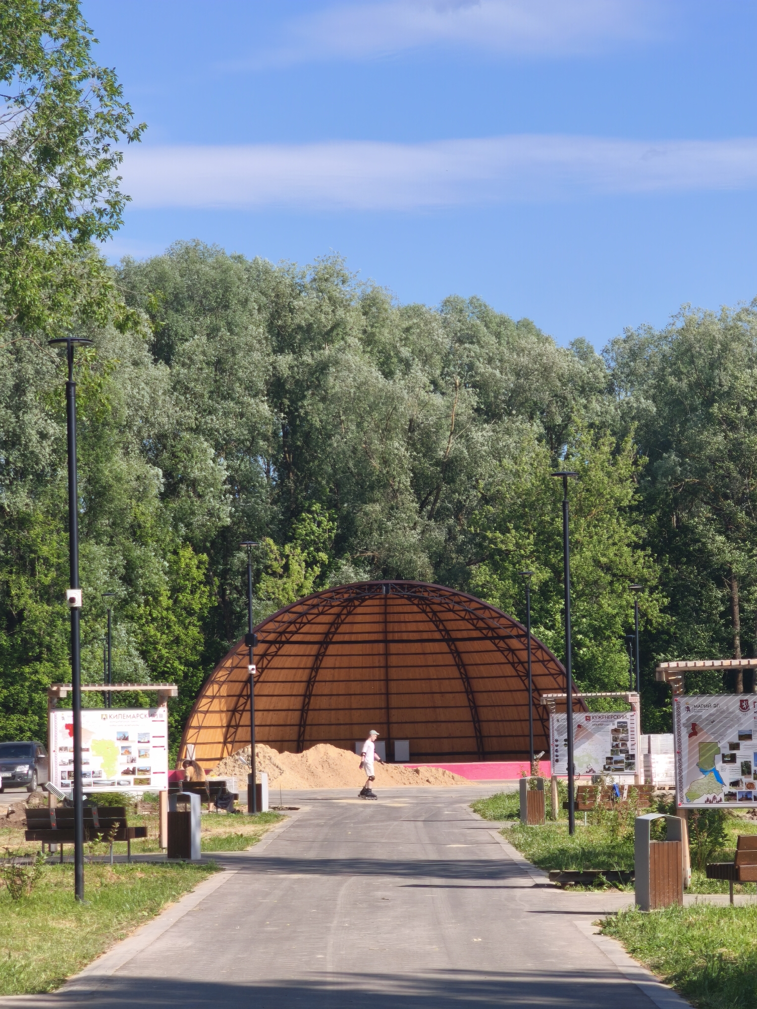 The image depicts a sunny day in a park in Yoshkar-Ola, Russia, with a clear blue sky and lush green trees forming the background. In the foreground, a paved pathway leads towards a large, dome-shaped structure with a visible wooden framework and an orange translucent covering. A person is seen rollerblading on the pathway. To the left and right of the pathway, information boards are displayed, and there are benches and trash receptacles. A car is parked on the left side of the frame, near one of the information boards. The area around the dome appears to be under construction, with a large pile of sand visible inside. The overall scene suggests a public outdoor space, possibly a recreational area or an event venue, during a warm day.