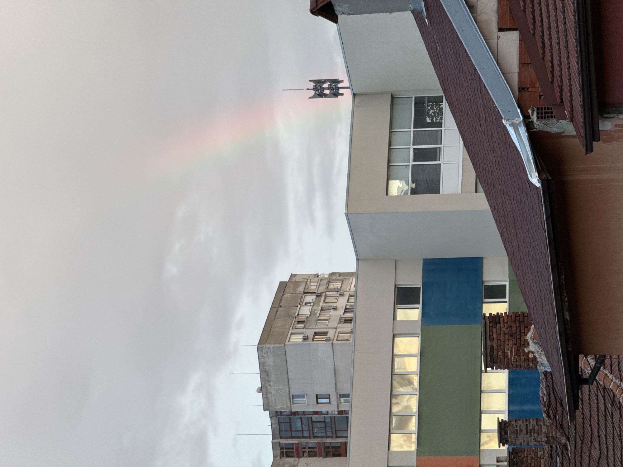 The image captures an overcast sky in Sofia, Bulgaria, with a faint rainbow appearing on the right side. In the foreground, several red-tiled rooftops are visible, some with brick chimneys. Behind the rooftops, a modern building with a light-colored facade is seen, featuring large windows that reflect the sky. Further in the background, a taller, older residential building with multiple windows and balconies stands. The overall scene is urban and residential, with a hint of natural phenomena in the form of the rainbow.