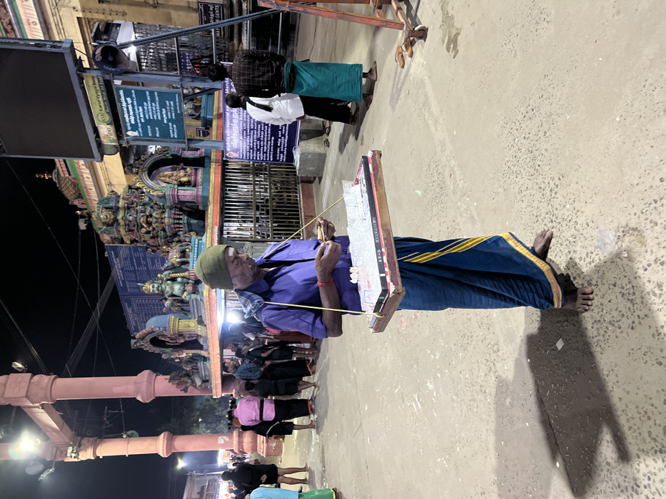 This nighttime image, captured in Palani, India, depicts an elderly man engaged in an activity outside a vibrant Hindu temple. The photograph is oriented sideways.
The main subject is an elderly man, seen in the foreground, wearing a purple long-sleeved shirt, a green or olive-colored cap, and a blue dhoti with a distinctive yellow border. He is barefoot and has a red thread or band on his right wrist. He holds a rectangular board, possibly a wooden slate, suspended by yellow strings around his neck or shoulders. With a thin, stick-like implement, he appears to be writing or drawing on the board, which has faint markings or text on its surface.
In the mid-ground and background, several other people are visible. Two individuals stand from behind, one in a white shirt and black trousers, the other in a green lungi and black shirt. Further back, a crowd gathers, some dressed in everyday attire, including one person in a pink top. The ground is a light-colored paved surface, and a pair of sandals lies discarded, suggesting entrance to a sacred area.
The setting is clearly a temple environment, characterized by an elaborately sculpted and brightly colored gopuram or temple entrance in the background. Intricate carvings of deities and mythological figures adorn the temple structures. Large, reddish-brown stone pillars stand prominently on the left side of the frame (when upright), possibly part of a mandapam or gateway. The scene is illuminated by artificial lights, casting shadows on the ground.
Visible text on a blue signboard in the mid-ground includes Tamil script, with partially readable English text below it indicating "WAY TO GENTS" and "WAY TO LADIES," likely directing pilgrims to amenities or specific queues. The overall atmosphere appears to be one of quiet devotion and activity, typical of a temple town at night.