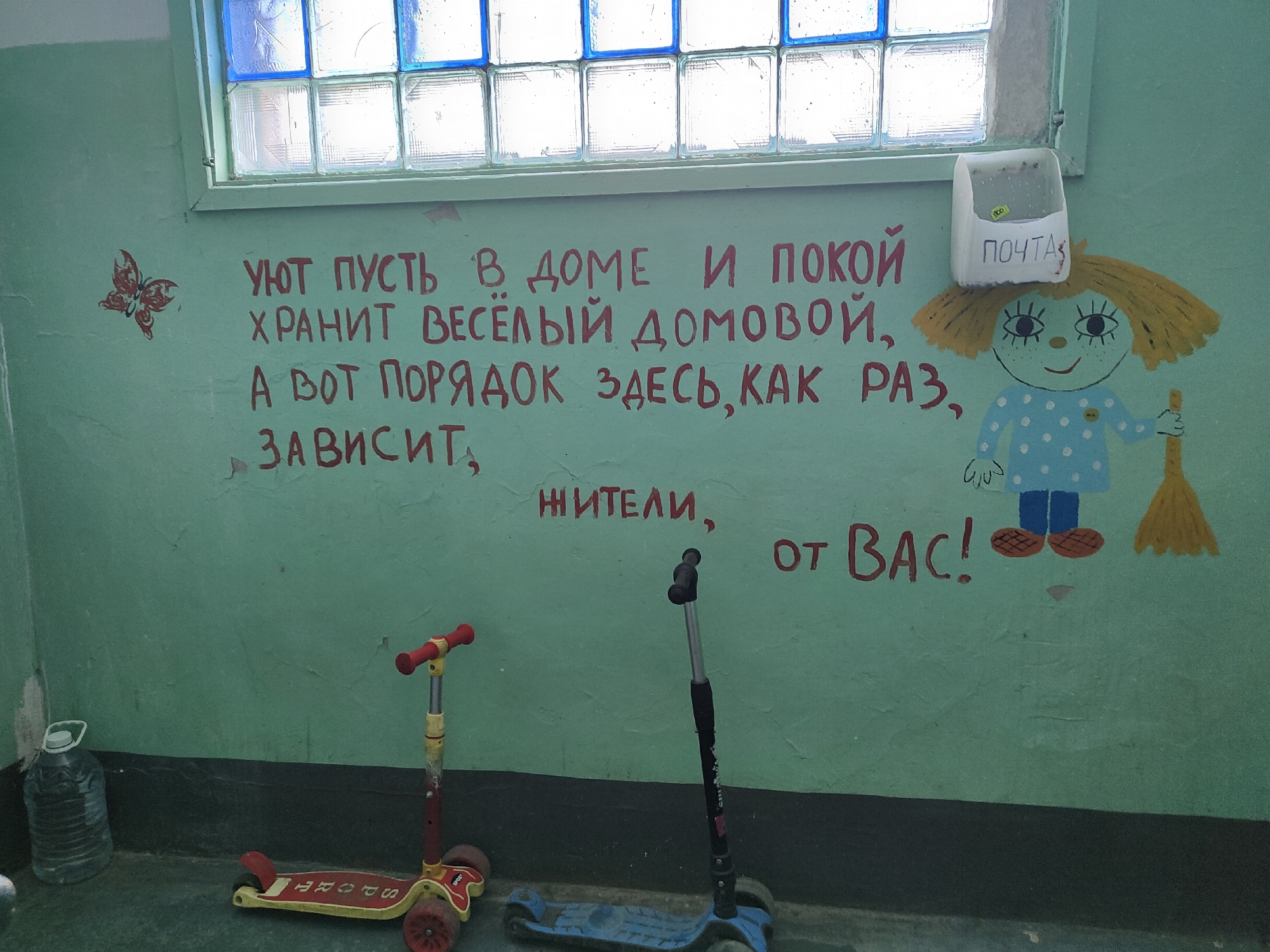 The image depicts an interior wall adorned with a mural and graffiti, located in Jaña Būqtyrma, Kazakhstan. The mural features a simplistic drawing of a girl holding a broom, with a mailbox labeled "ПОЧТА" (POST) positioned above her head. Red graffiti across the wall translates to "Let coziness and peace be kept by a cheerful house spirit, but order here, as it depends on the residents!" and "from YOU!". A small red butterfly stencil is visible to the left of the text. In the foreground, two scooters, one red and yellow, and another blue and black, are placed on the floor. A large plastic water bottle is situated to the left of the scooters. The wall is painted a light green color, and a window with frosted glass blocks is visible at the top of the frame. The overall environment appears to be a functional space, possibly a common area or hallway.