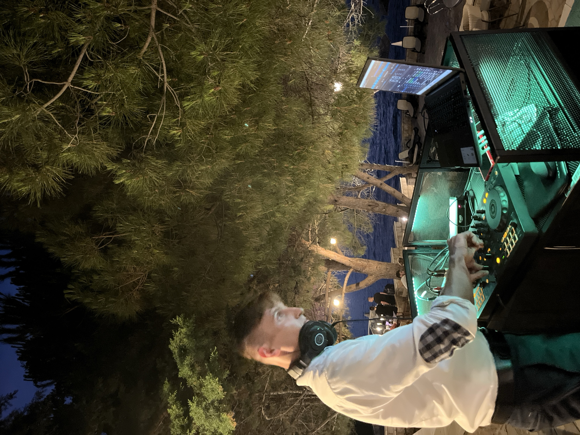 A young man wearing a white collared shirt and headphones around his neck is operating a DJ console outdoors at night. The DJ console is illuminated with green lights and includes a laptop, turntables, and various controls. The man's right hand is on the console, suggesting he is actively mixing music. In the background, palm trees and other foliage are visible, silhouetted against a dark sky. Beyond the trees, a body of water, likely the Adriatic Sea, can be seen with gentle waves. The scene appears to be a beach or waterfront venue, possibly in or near Pečurice, Montenegro, given the presence of lounge chairs and tables set up for an event. The overall ambiance is relaxed and festive, with the subtle lighting creating a moody atmosphere.