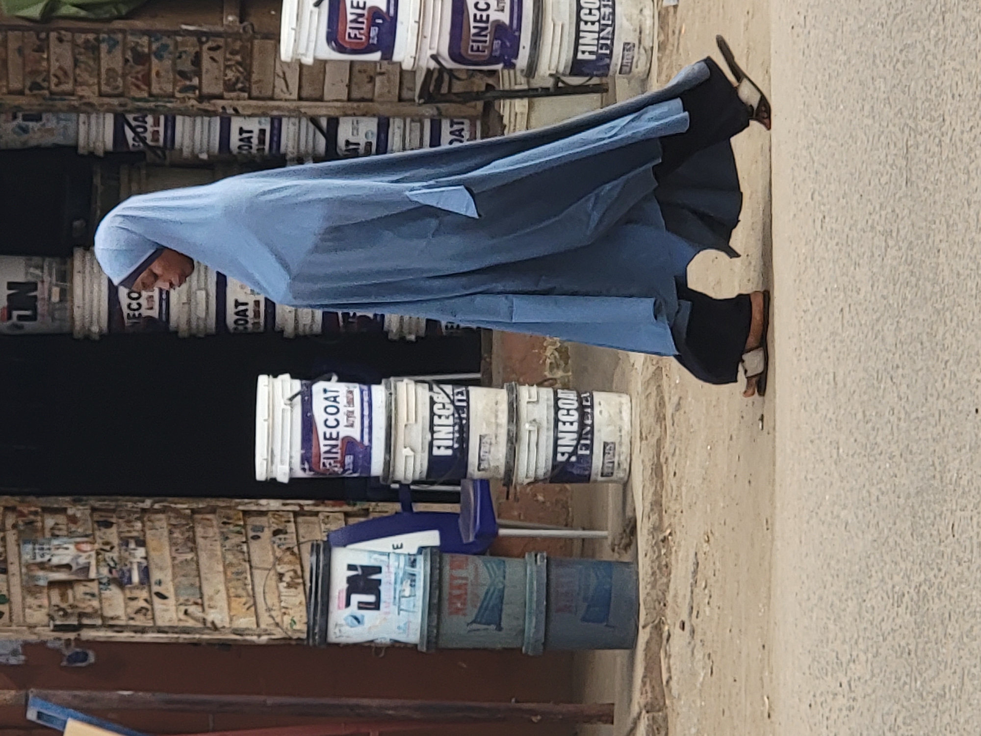 A woman wearing a blue head covering and robe walks past a stack of paint buckets in Maiburiji, Nigeria. The buckets are stacked against a weathered wall or display shelf. Some buckets feature the brand "FINECOAT." The road or sidewalk appears dusty or unpaved.