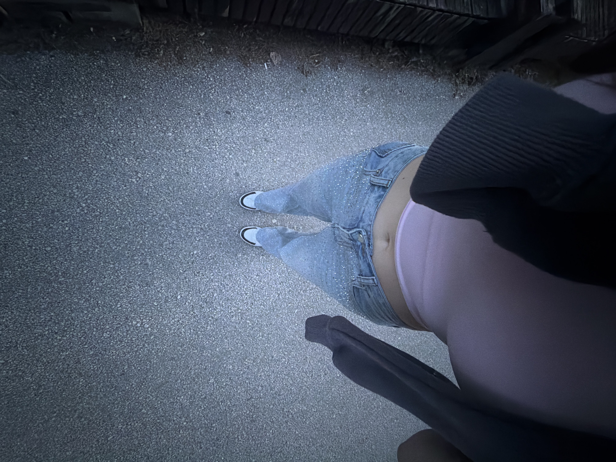 A high-angle shot looking down at a person standing on an asphalt surface. The person is wearing light blue jeans adorned with rhinestones, white sneakers with black trim, and a pink undergarment visible beneath the jeans' waistband. A dark-colored garment, possibly a jacket or sweater, is partially visible on the right side of the frame. The asphalt appears to be a pathway or street surface. At the top of the frame, a wooden structure, potentially a fence or building facade, is seen. The lighting suggests it is either dusk or night. There are no visible landmarks to determine the specific location within Pazardzhik, Bulgaria.