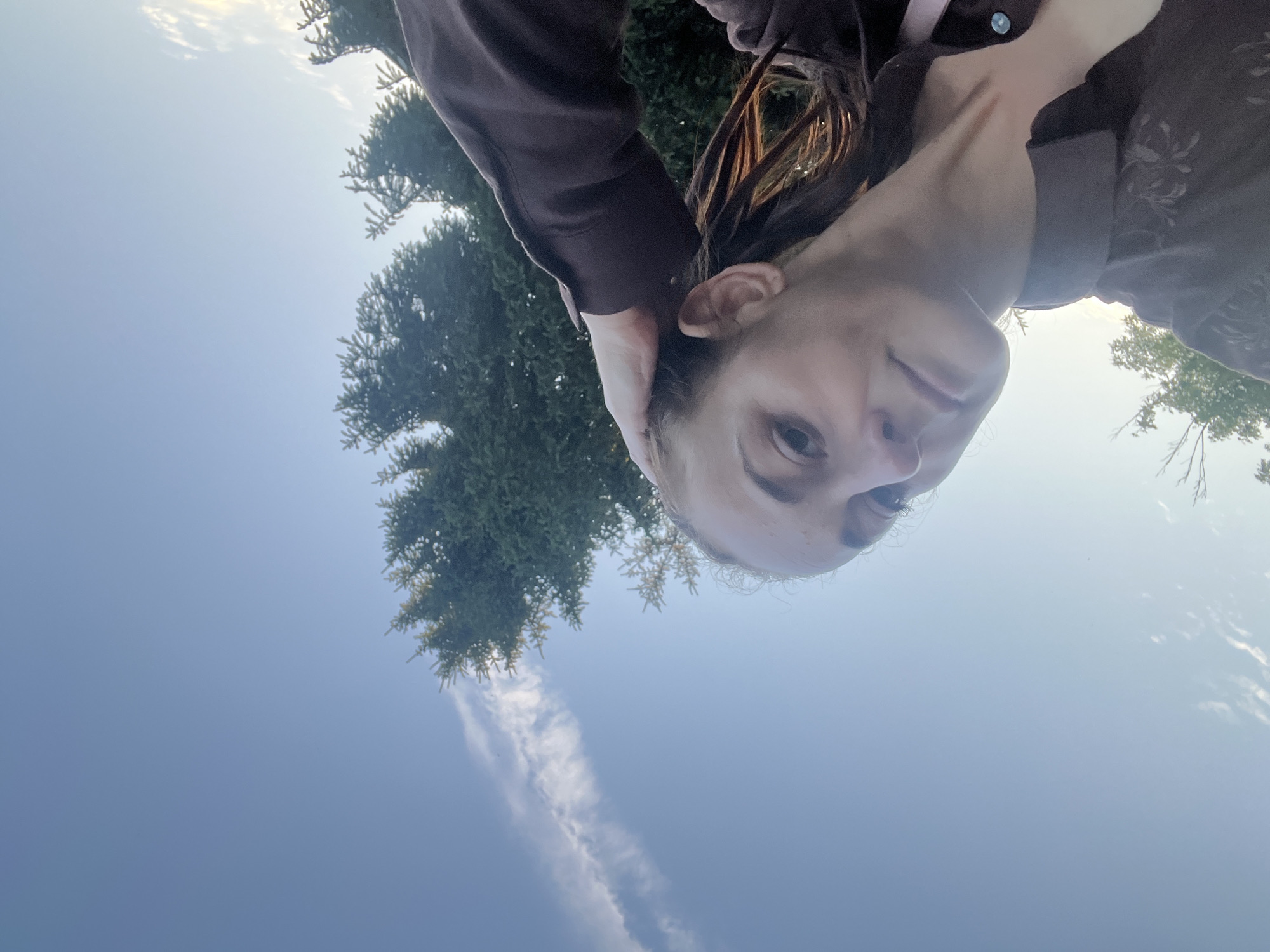 A woman is seen from a low angle, holding her right hand to the side of her head. She is wearing a dark brown collared shirt. The background is a clear sky with a wispy cloud formation and the upper portion of a dark green coniferous tree. The lighting suggests it is daytime, possibly late afternoon or early evening.