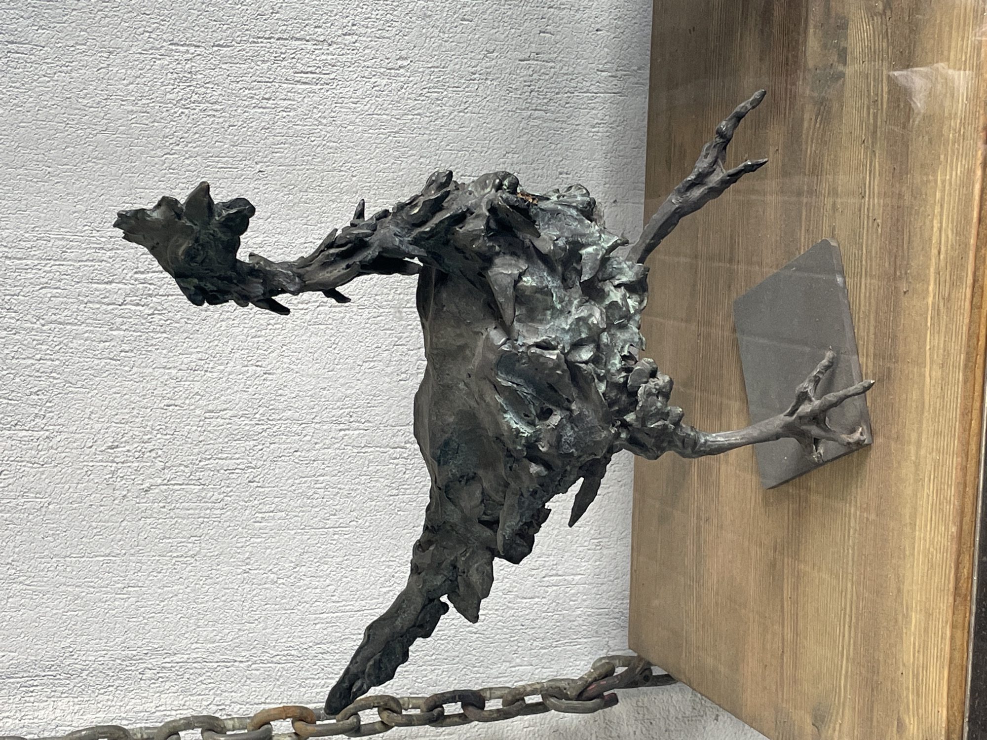 The image shows a metal sculpture of a fantastical bird-like creature mounted on a wooden board against a rough, white textured wall. A chain is visible below the sculpture. The context or environment suggests it could be an exhibit in a museum or gallery. It's likely this photo was taken in Saint Petersburg, Russia, based on the presence of similar artwork in various collections and exhibitions there.