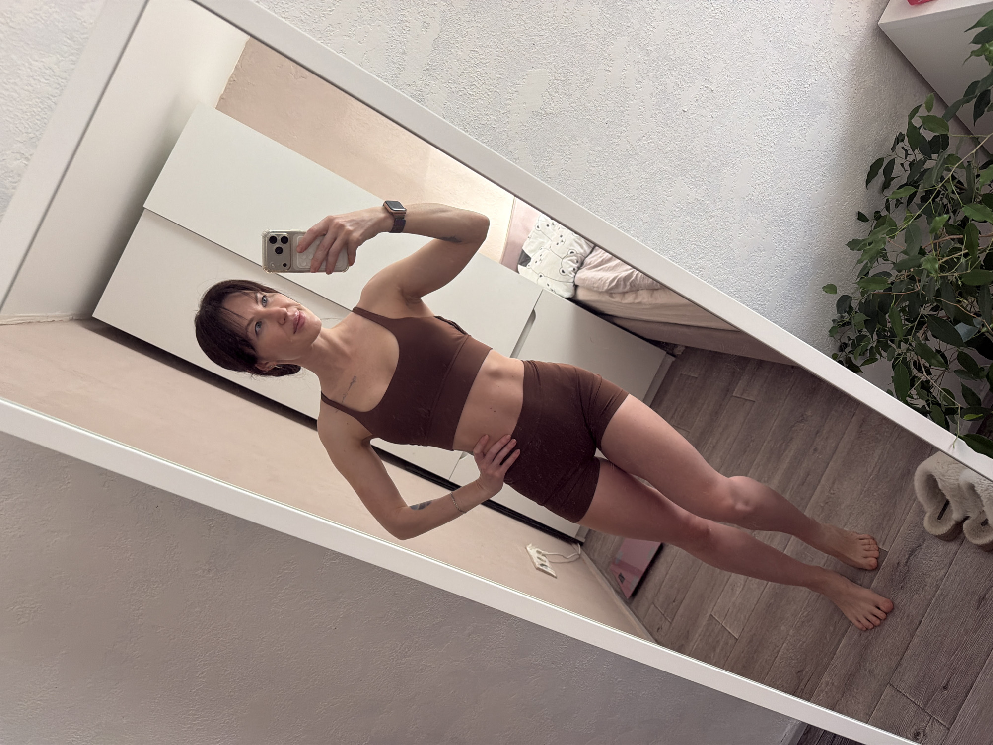 A woman stands in front of a large, white-framed mirror, taking a selfie with her iPhone. She is wearing a brown sports bra and matching shorts. Her left hand is on her hip. The mirror reflects a bedroom scene, including a bed with patterned bedding, a white wardrobe, and a plant in the corner. The flooring appears to be wood-look planks. The location may be in Perm, Russia, but no direct evidence indicates this.