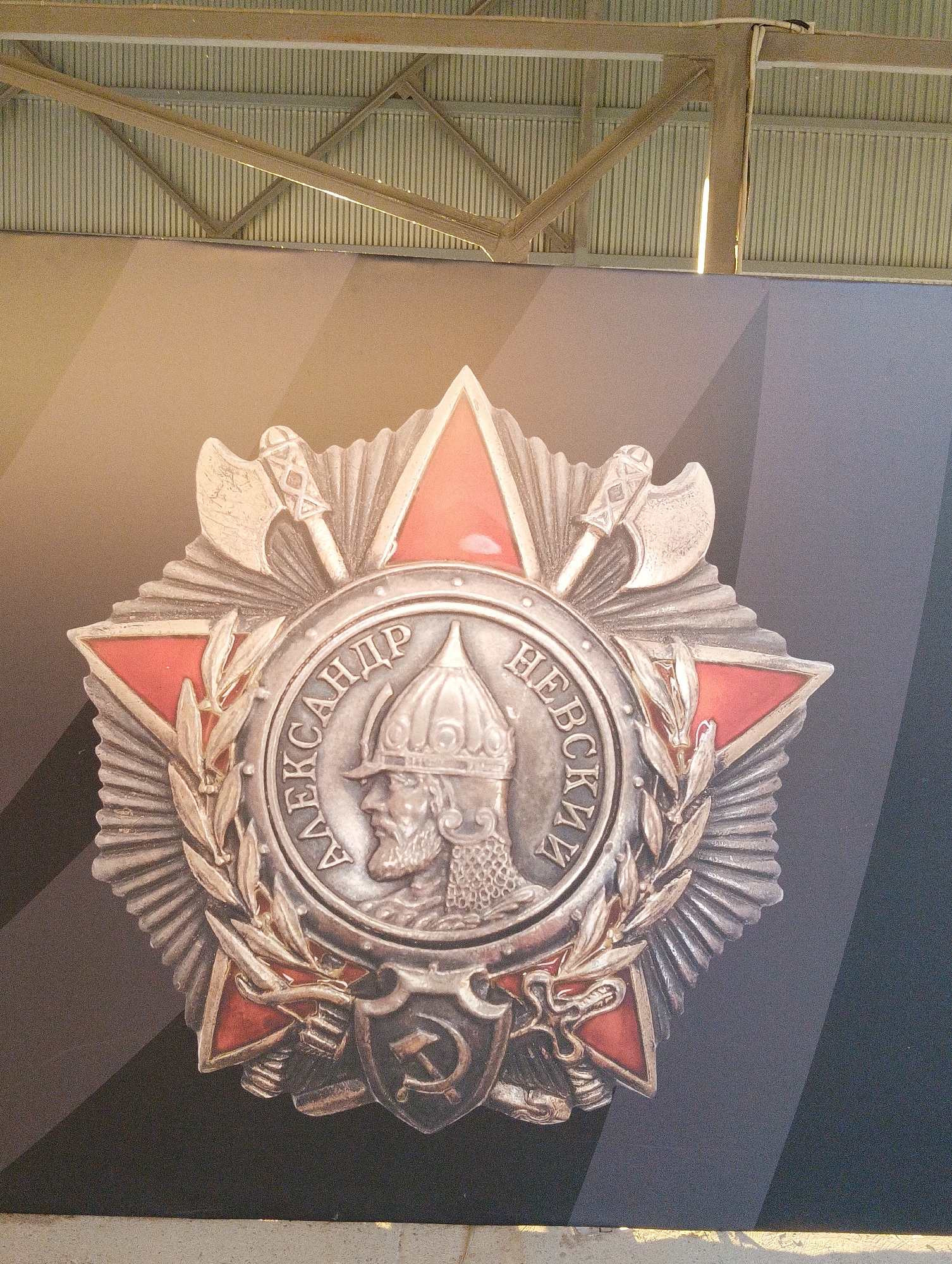 The image shows a close-up of a large medal featuring a profile of a man in a helmet, surrounded by laurel branches and crossed axes. The medal is predominantly silver with red accents. Cyrillic lettering is visible around the central portrait. Below the medal is a shield with a hammer and sickle emblem. The medal is displayed against a dark background with a geometric pattern of light and dark gray stripes. Above the medal, a corrugated metal ceiling and industrial metal beams are visible, suggesting an indoor setting. The overall impression is of a museum exhibit or public display. The medal is likely the Order of Alexander Nevsky, a Soviet and Russian award. The image itself does not contain any visual information to pinpoint its location in Pesochny, Russia.