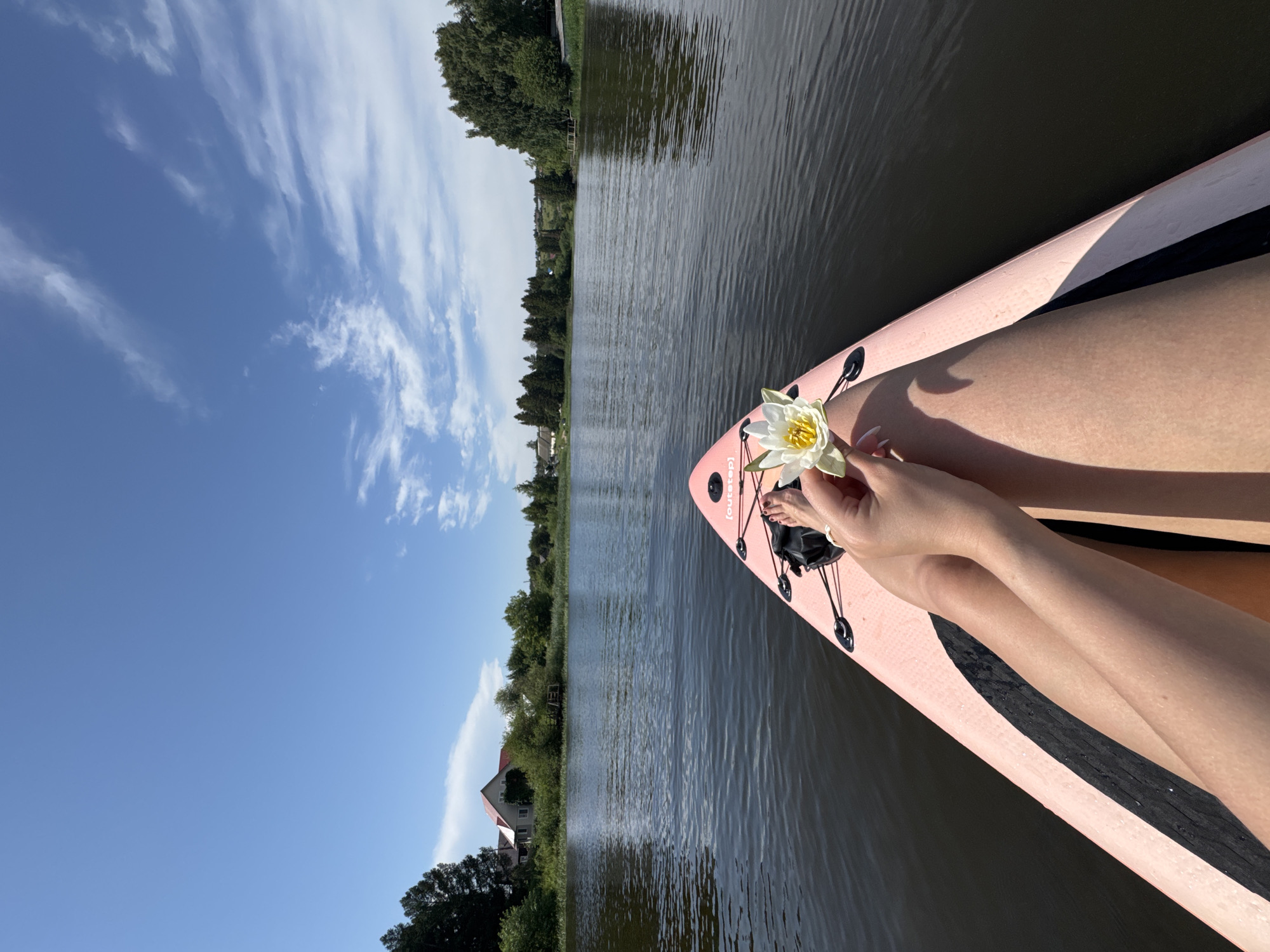 A person sits on a pink paddleboard on a body of water, holding a white water lily. The person's legs and feet are visible in the foreground. The paddleboard has "outstep" written on it. The water is dark and calm, with a slight ripple on the surface. In the background, there is a lush green shoreline with trees and houses. The sky is blue with scattered white clouds. The location is Chastyye, Russia.