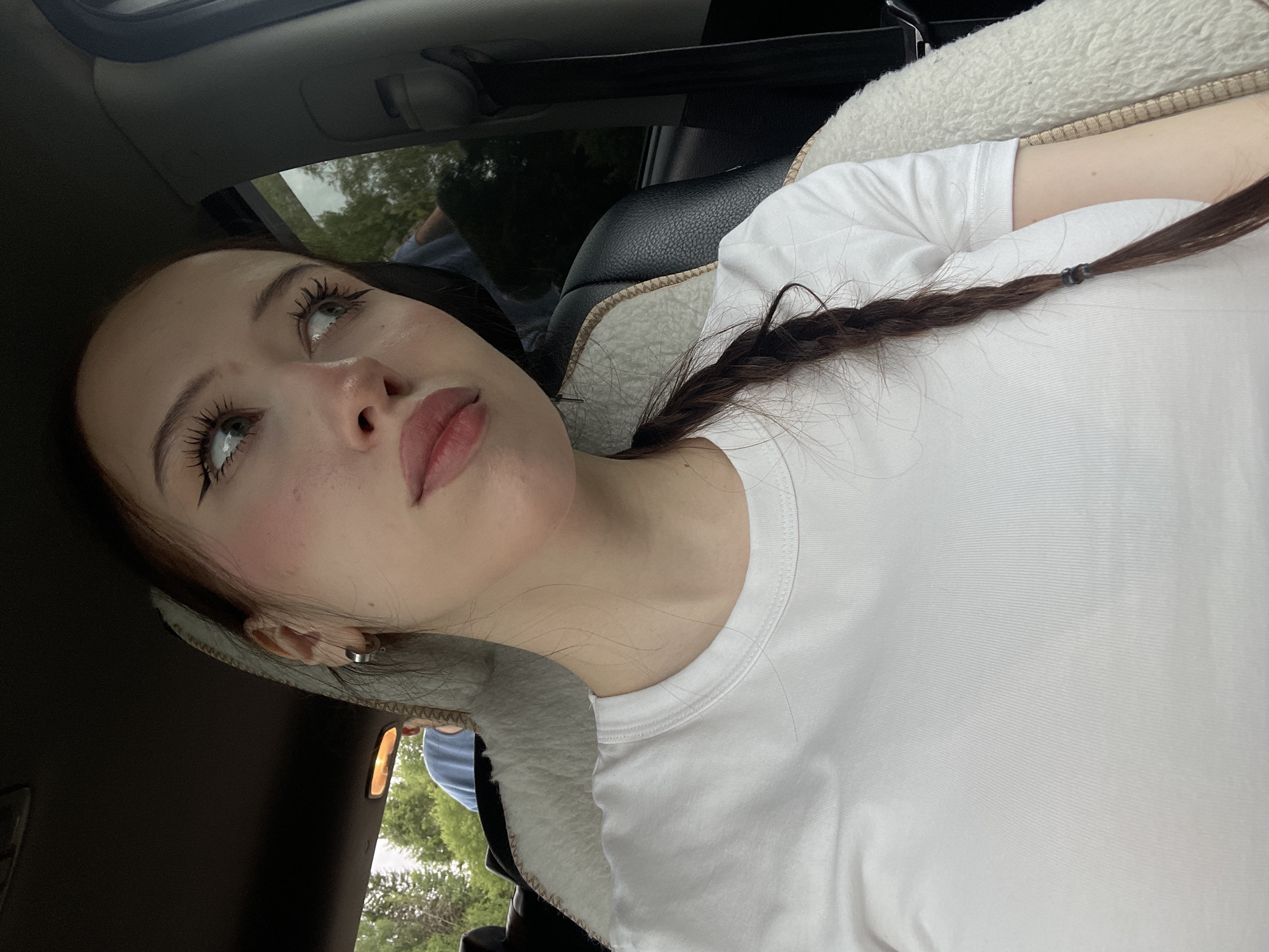 A young woman with long, dark hair braided into a single plait is seated in the passenger seat of a car. She is wearing a white t-shirt and has makeup on, including winged eyeliner and blush. Her gaze is directed to the right, away from the camera. In the background, through the car window, trees and a cloudy sky are visible. Another person, wearing a blue shirt, is visible in the back seat. The image appears to have been taken during the daytime.