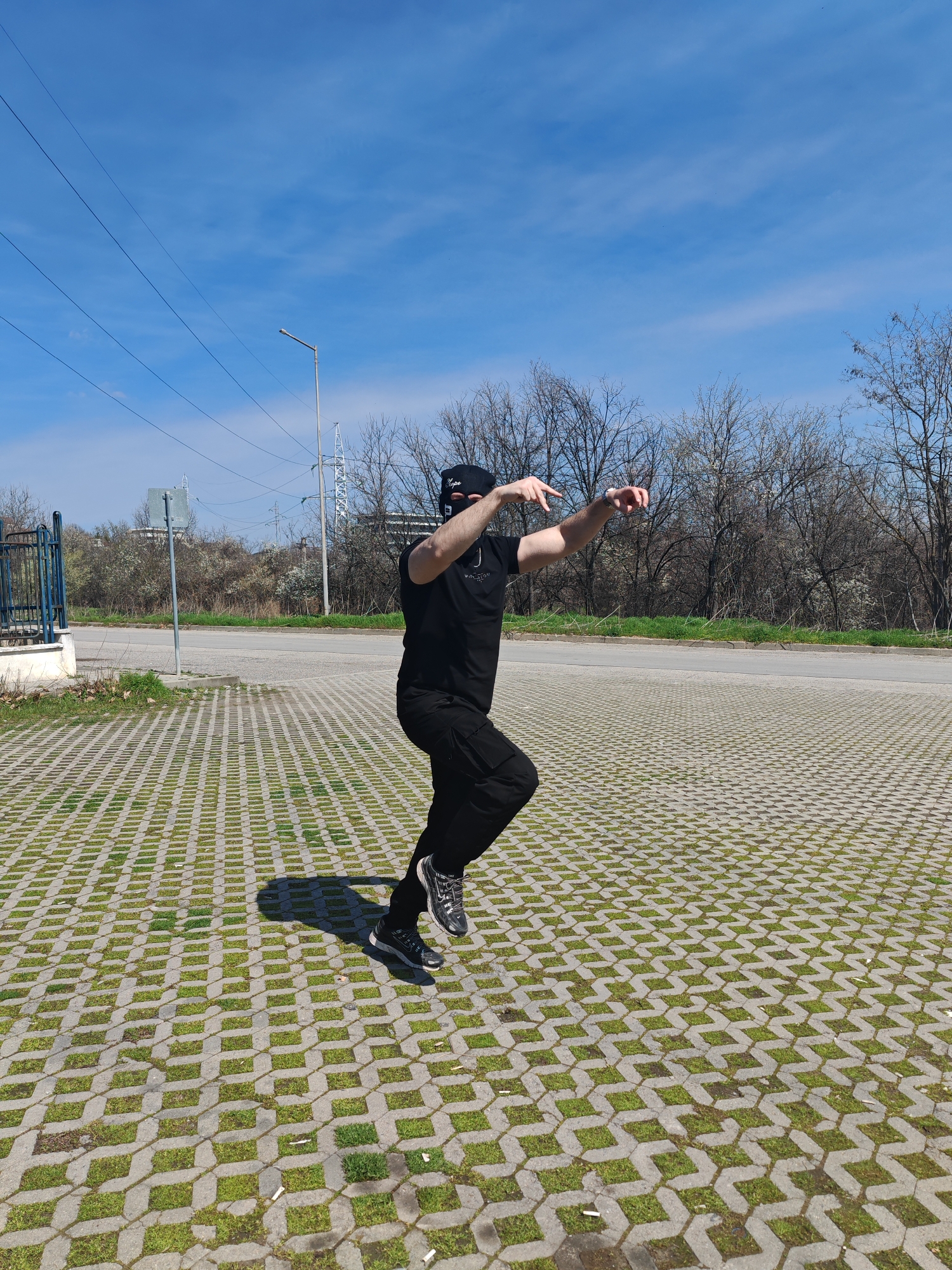 A single individual, dressed entirely in black attire including a t-shirt, cargo pants, athletic shoes, and a black balaclava with "Hope" inscribed in white, is depicted in mid-air on a patterned concrete paver surface. The person's right leg is bent with the foot off the ground, and the left leg is extended backward, also airborne. Both arms are outstretched forward and slightly upward, with open hands and spread fingers. The subject casts a distinct shadow to their left on the grass paver ground, which contains scattered cigarette butts. To the left, a blue metal fence and a gray signpost are visible. An asphalt road traverses the mid-ground, beyond which lies a strip of green grass and a line of bare deciduous trees. The background includes multiple utility poles, power lines, a streetlamp, and a metal lattice transmission tower, with a faint building in the far distance. The scene is illuminated by direct sunlight under a clear blue sky, located in an urban-edge environment in Shumen, Bulgaria.