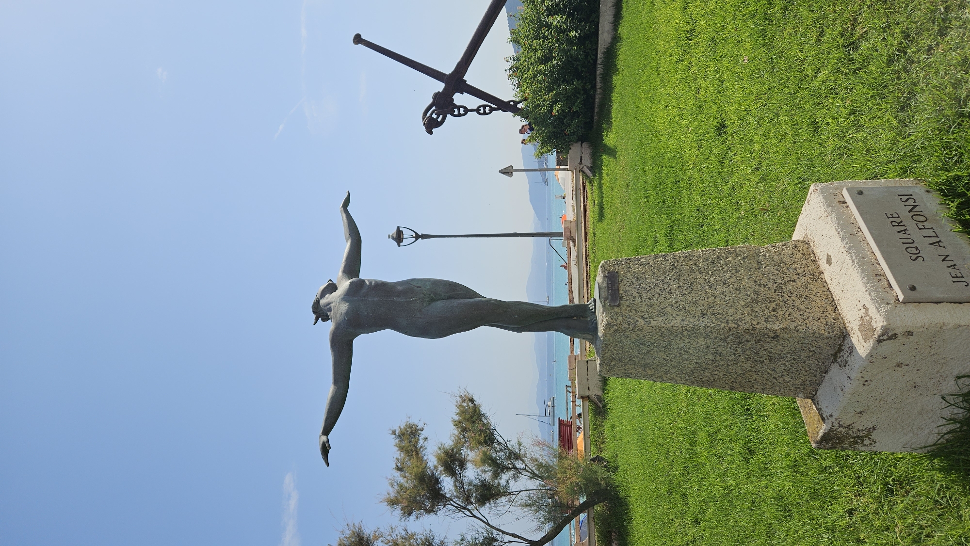 A bronze statue of a female figure with arms outstretched stands on a granite pedestal in L'Île-Rousse, France. The statue is located in a grassy area next to the sea, with a beach and boats visible in the background. An anchor is also visible in the background. The base of the pedestal has a plaque that reads "SQUARE JEAN ALFOnSI." The statue faces away from the camera, towards the sea. The sky is blue and clear.