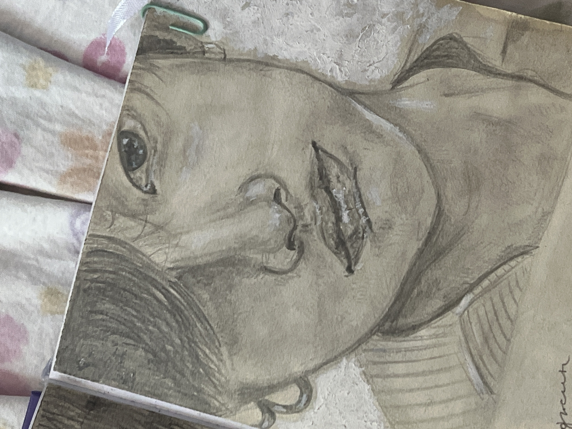 The image displays a close-up, sepia-toned watercolor painting of a person's face, rendered in a sketchbook. The face is positioned centrally, with the right side of the face more prominently visible. The person has dark hair styled to partially cover the left side of their face, revealing a portion of their forehead and right eye. The eye is looking directly at the viewer, with detail captured in the iris and pupil. The nose is depicted with subtle shading, and the lips are slightly parted, showing some texture and highlights. The person is wearing a collared garment, suggested by horizontal lines on the chest area. The background of the painting appears textured with light and dark patches. A green paperclip is attached to the right edge of the sketchbook page. The sketchbook itself is open, and the surface beneath it is a light-colored fabric with scattered floral or circular patterns in pink and light yellow. The bottom left corner of the painting features a handwritten signature or inscription in dark ink. There is no visible indication of the city of Shymkent, Kazakhstan, in the image.