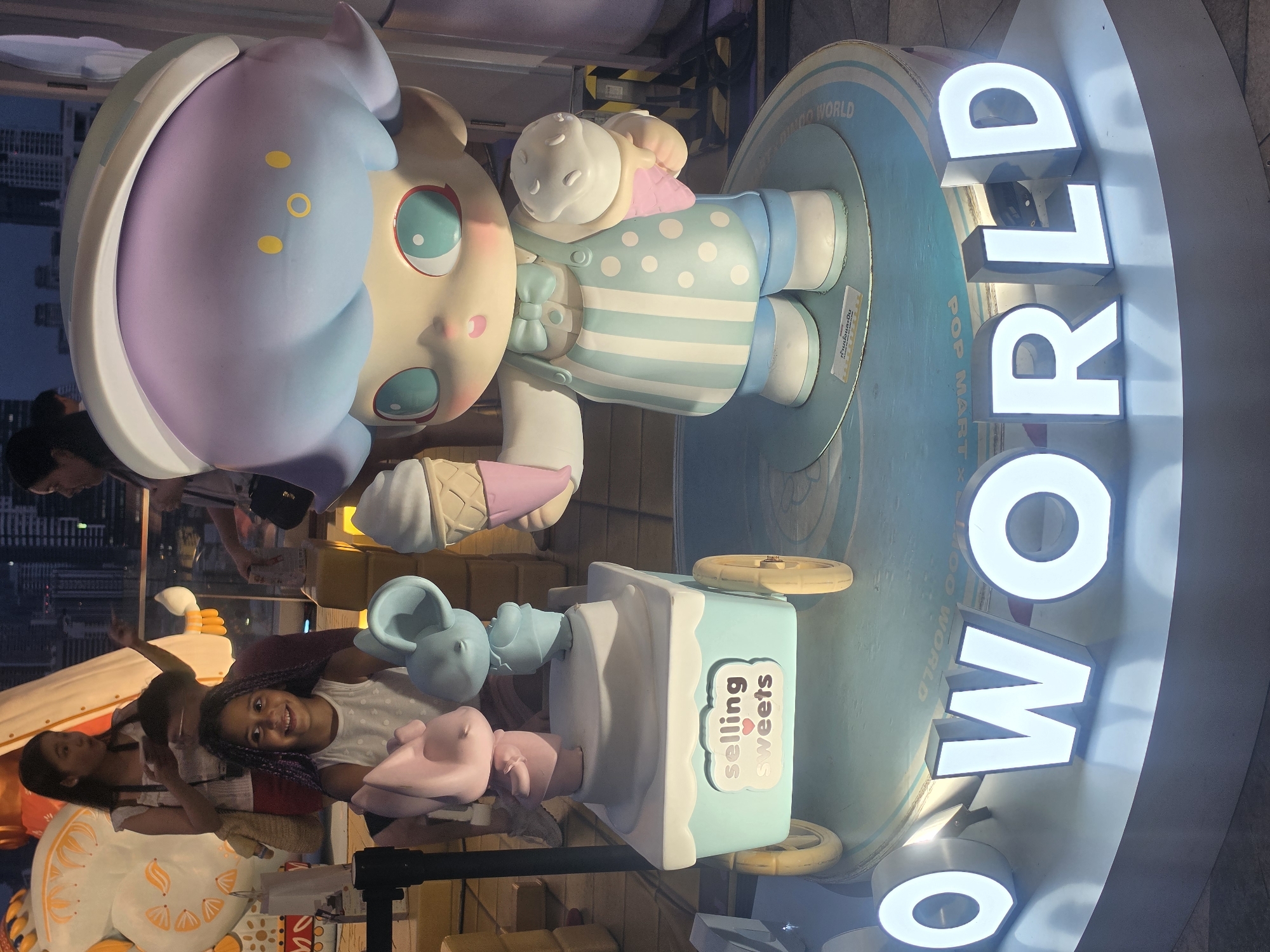 The image shows a large, colorful statue of a cartoon character, possibly a bear, with elements like an ice cream cone and a "selling sweets" cart. The statue sits atop a round platform with the text "POP MART DIMO WORLD" and "O WORLD" in large, illuminated white letters. Two young people are visible in the foreground, appearing to interact with or pose with the statue. The surroundings suggest an outdoor location in Bangkok, Thailand, as indicated by the Thai text on the signage. The scene is brightly lit and likely located in a commercial or public area where such displays are common.