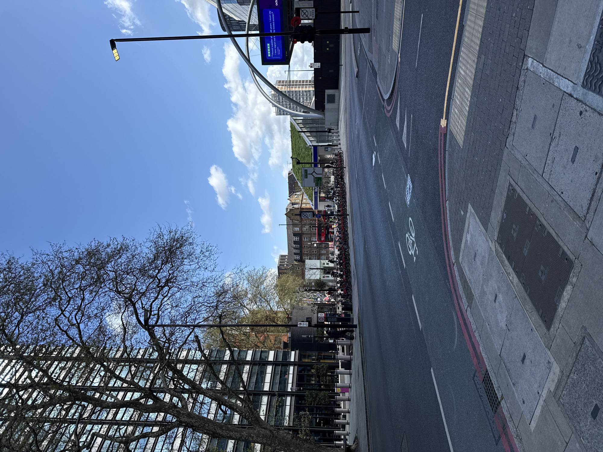 The image depicts a street scene in London, United Kingdom, on a sunny day. The perspective is a slightly elevated street-level view looking down the length of the road.
On the right side of the road, there's a pedestrian walkway with a textured crossing area. The road itself has a designated bicycle lane indicated by a white bicycle symbol painted on the surface. A double red line marks the edge of the carriageway.
Along the left side of the street, there's a modern glass-paneled building fronted by a row of metal bollards. A tree with bare branches stands in front of this building. Further along, there is a line of parked bikes and a bus station.
In the background, there are buildings, trees, and a pedestrian bridge structure with advertising on it that reads 'Octopus'. The sky is blue with scattered white clouds. A lamppost extends from the left side of the street into the sky. The traffic light is visible in the background, on the right side.