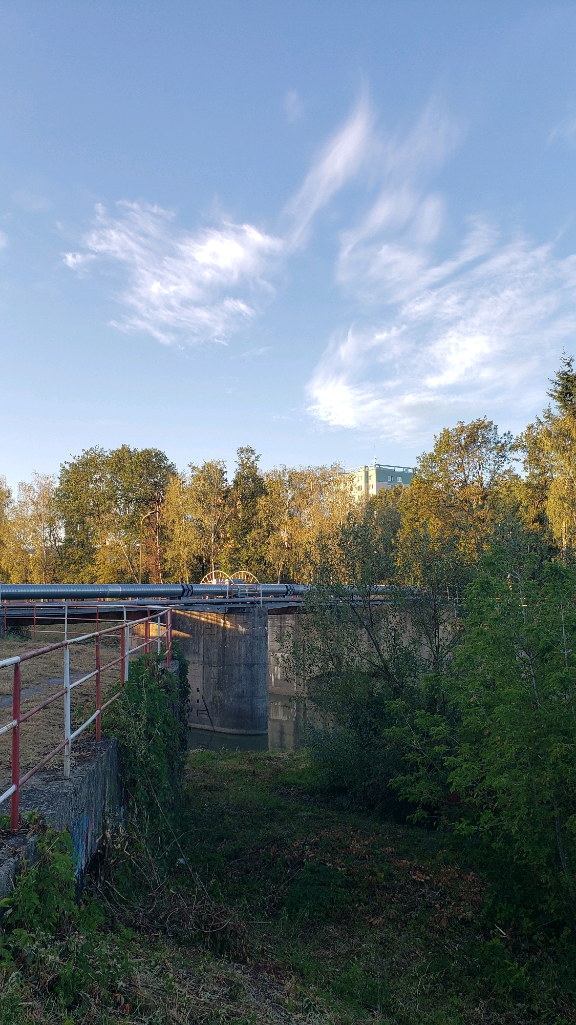 The image shows a view of a large pipe supported by concrete structures, likely part of an industrial or utility infrastructure. The pipe runs horizontally across the frame, elevated above a body of water. The surroundings are characterized by lush green vegetation, including trees and bushes, suggesting an outdoor setting. In the background, a multi-story building, possibly an apartment complex or office building, is visible amidst the trees. The sky is a clear blue with wispy white clouds, indicating fair weather. The scene appears to be a section of the city of Prešov, Slovakia, possibly near the Sekčov district, where such infrastructure and water bodies are found. There are no people or animals visible in the image.