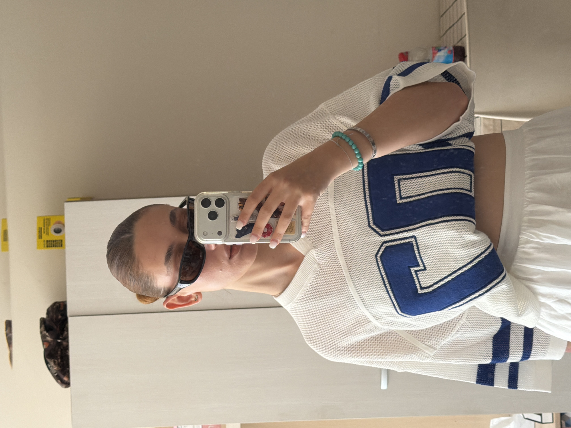 A woman is taking a selfie in a mirror indoors. She is wearing a white crop top with blue accents, a white skirt, and sunglasses. She has two bracelets on her left wrist. The background appears to be a room with light-colored walls and furniture. The location is not identifiable as Sofia, Bulgaria, based on the visual information provided.