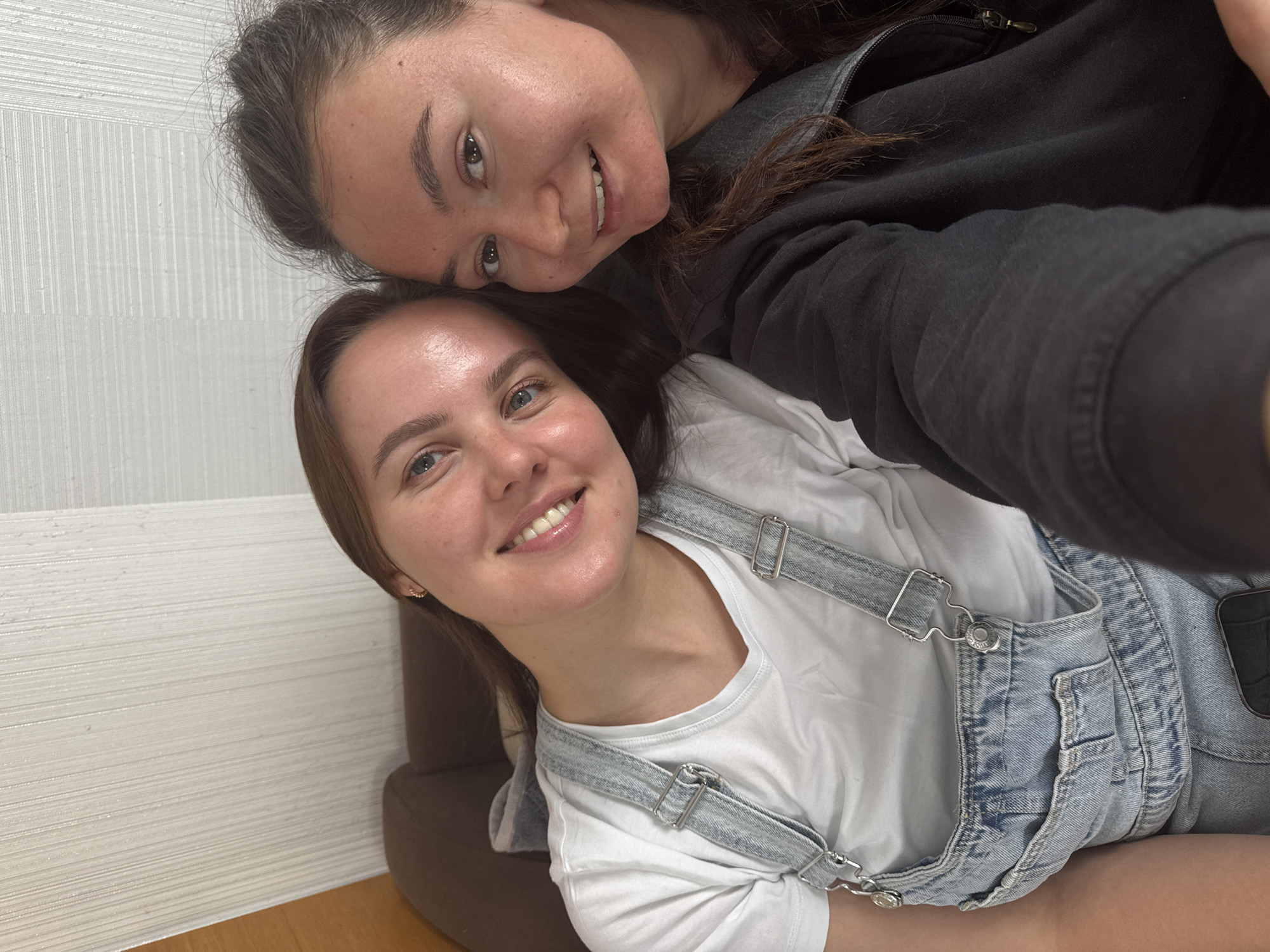 Two women are posing closely for a selfie. The woman on the bottom is wearing a white t-shirt and blue denim overalls. The woman on top is wearing a black zip-up hoodie. The location appears to be indoors, near a brown couch. It is not possible to determine if this image was taken in the city of Дойная, Russia.