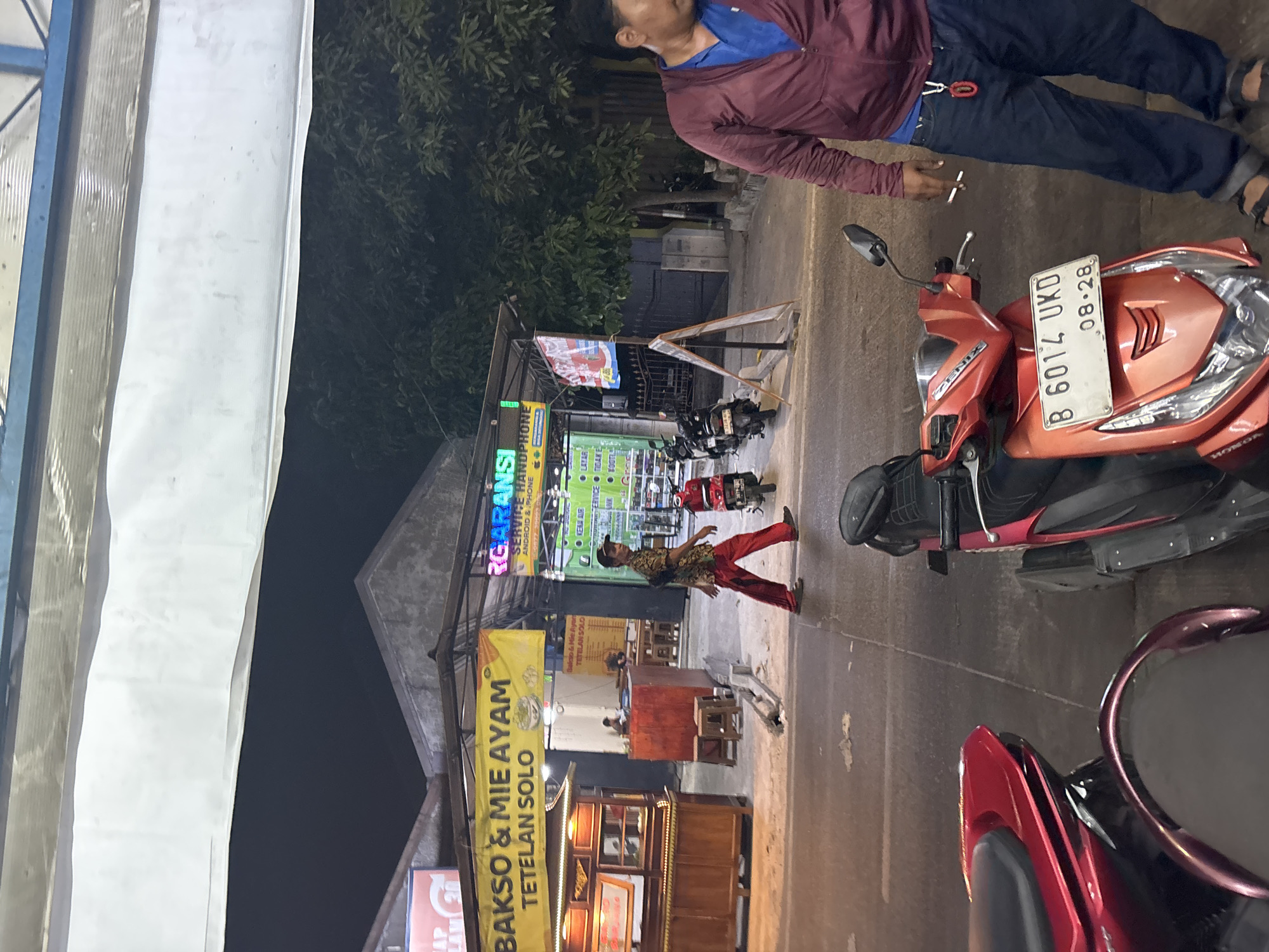 The image depicts a street scene at night in North Jakarta, Indonesia, likely near a commercial area with food stalls and shops. In the foreground, several motorbikes are parked. To the left, a red motorbike is partially visible. To the right, an orange motorbike is prominent, bearing the license plate "B 6014 UKD 08.28". A man in a maroon jacket and blue shirt, wearing dark pants and sandals, stands next to the orange motorbike, holding a cigarette.
In the mid-ground, a man wearing a patterned shirt and red pants walks across the street. Behind him, a food stall with a wooden counter and display cases is set up. A yellow banner above the stall reads "BAKSO & MIE AYAM TETELAN SOLO", indicating it sells beefball and chicken noodles from Solo. Further back, a green and white banner for "RG ARANSI" is visible, suggesting a cellphone repair or sales business. Another smaller banner with Indonesian text is also present.
The background shows a building with a metal roof and trees. The overall lighting suggests it is nighttime, with artificial lights illuminating the area. The environment appears to be an urban street with pedestrian and vehicle traffic.