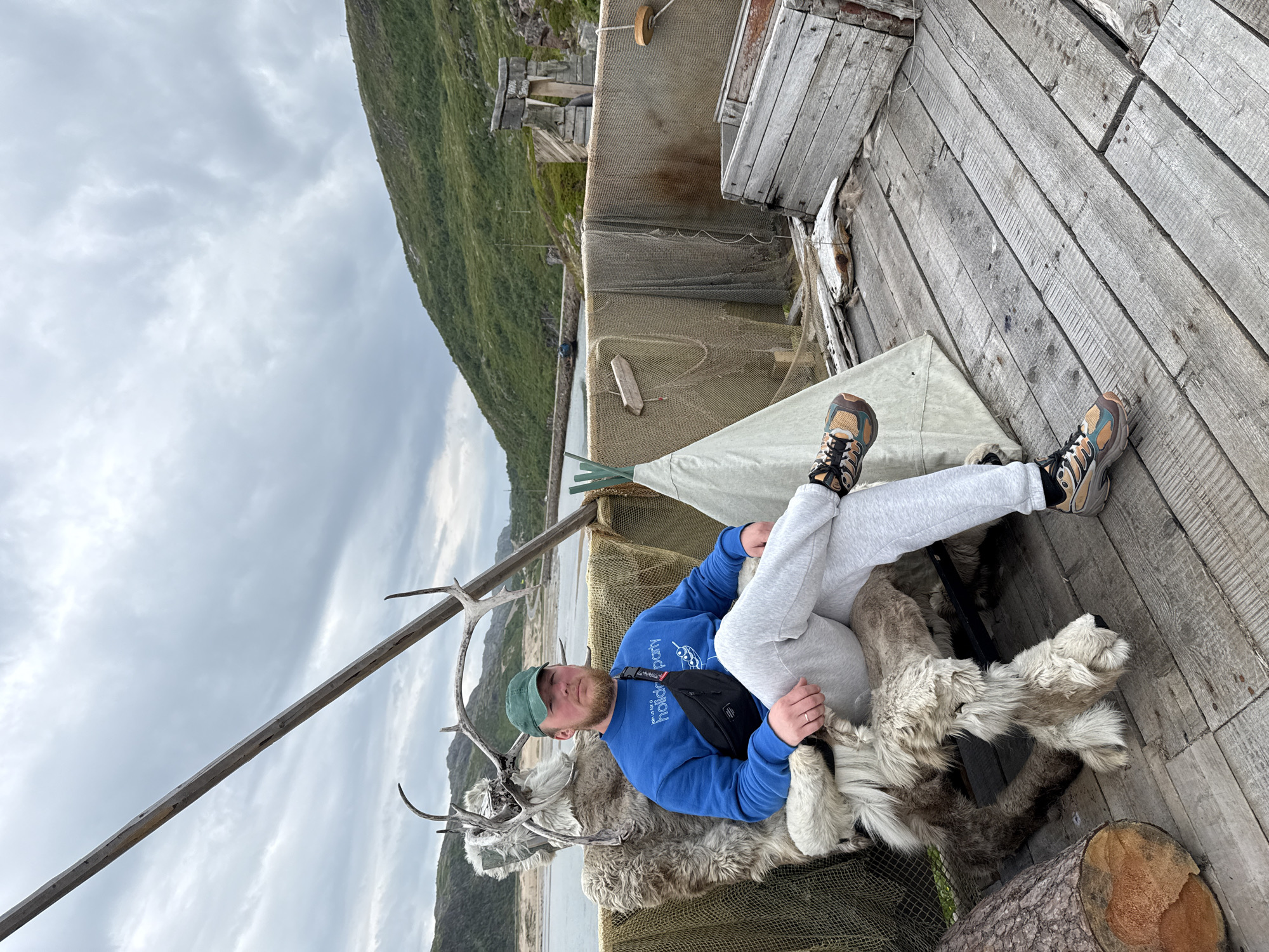 In Teriberka, Russia, a man with a beard wears a green cap, a blue sweatshirt with "join us for a holiday party" printed on it, grey sweatpants, and multi-colored athletic shoes. He is seated on a chair covered in reindeer hide, adorned with large antlers. The man has a black fanny pack slung across his chest. He is positioned on a wooden dock, with his legs crossed. To his right, a small, light-colored conical tent stands nearby. Behind him, fishing nets hang from a structure, and a wooden crate sits to the right. The background features a body of water, a distant green, rocky hill, and an overcast sky.