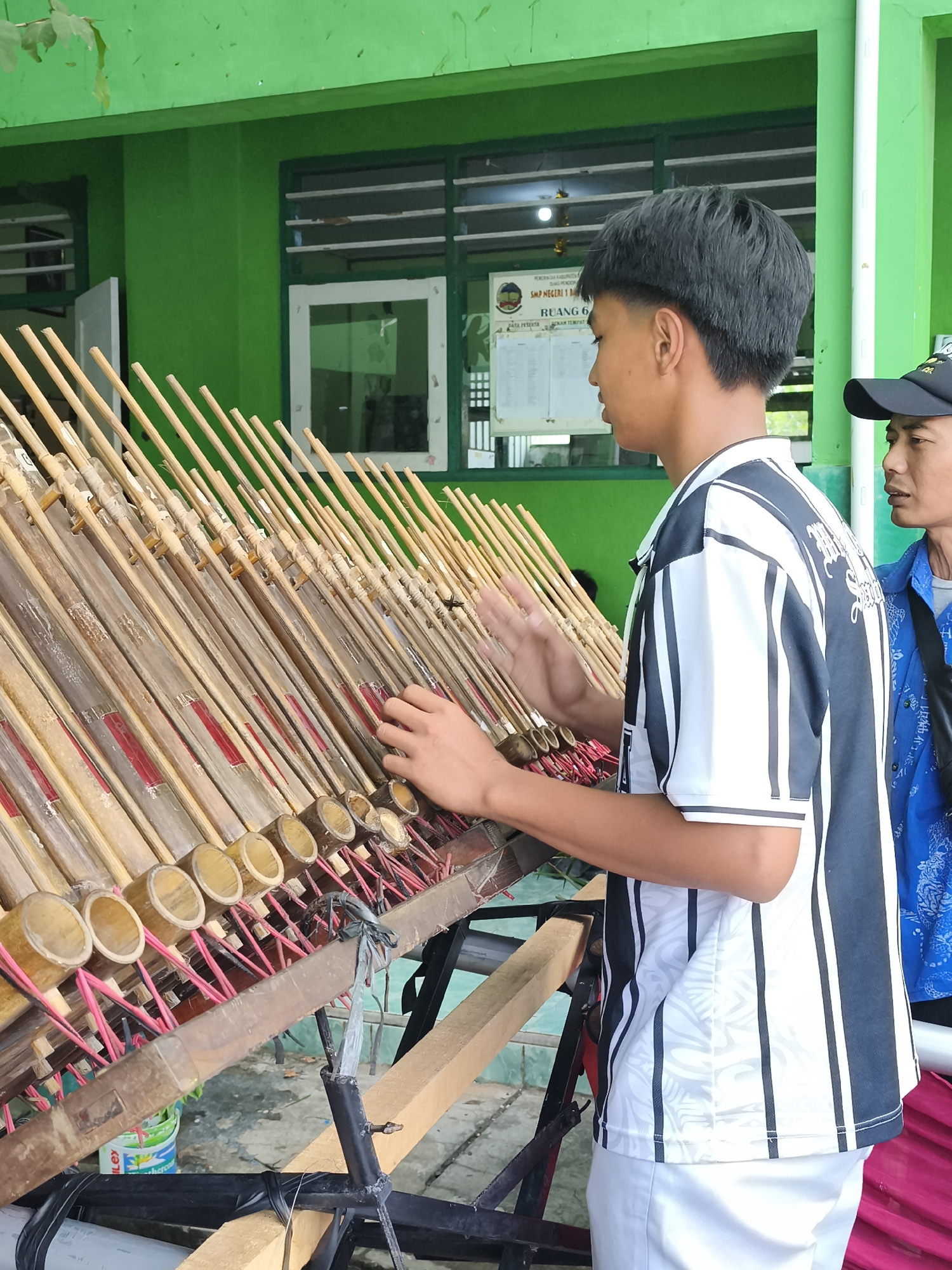 The image depicts two males in front of a traditional Indonesian musical instrument, likely an angklung, arranged in multiple rows. The instrument is made of bamboo tubes of varying lengths, suspended by pink strings and attached to a wooden frame. The foreground shows a young male with dark, short hair, wearing a black and white striped shirt and white pants, actively playing the instrument with his hands. To his right, another male, wearing a black cap and a blue patterned shirt, observes. The background features a green building with windows and a bulletin board displaying information in Indonesian. The setting appears to be outdoors, with some foliage visible at the top left. The overall scene suggests a cultural or educational event in Banyumas, Indonesia, possibly at a school, given the presence of the bulletin board with school-related text.